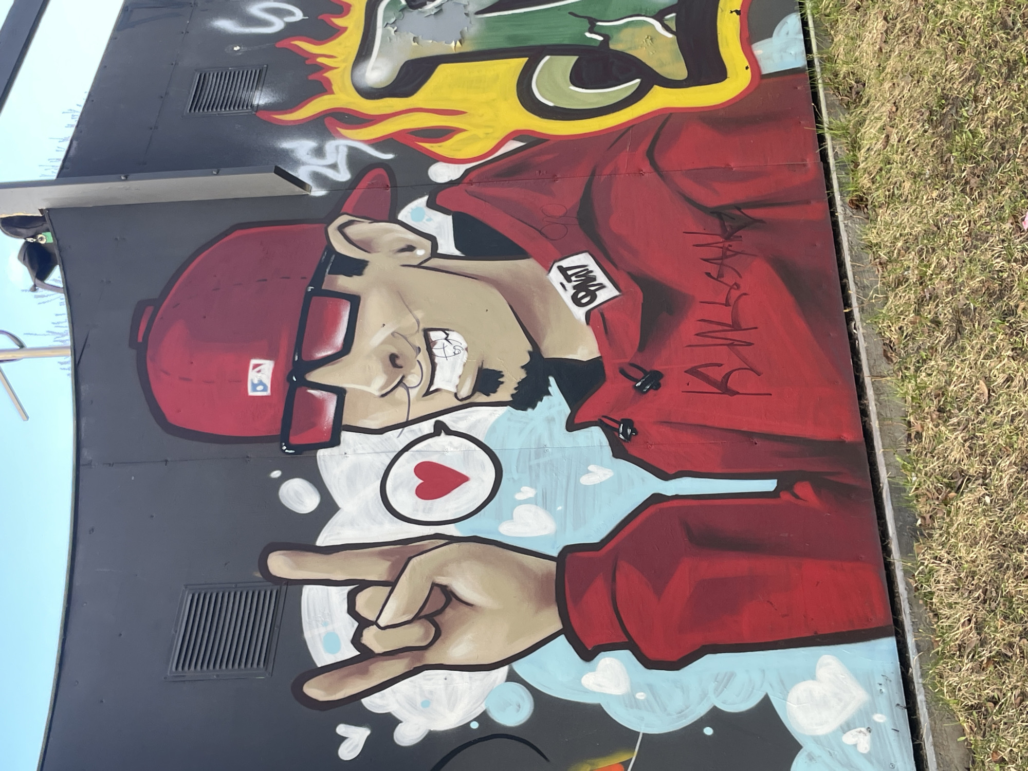 This image captures a vibrant graffiti mural on a dark grey outdoor surface, likely a utility box or wall, located in Izhevsk, Russia. The central subject is a stylized male character depicted from the waist up, rendered in a cartoonish style with bold outlines.
The character wears a red baseball cap featuring a simplified white "LA" logo, a red hooded sweatshirt, and dark-rimmed glasses. He has light skin, a dark beard, and a wide, cheerful smile that shows his teeth. His right hand (viewer's left) is shown making a "rock on" hand gesture, with index and pinky fingers extended. A white speech bubble containing a red heart floats near his head, indicating affection or positive sentiment.
The mural's background elements include yellow and red flame-like shapes at the top, transitioning to light blue cloud shapes interspersed with white hearts and bubbles towards the bottom. Visible text on the character's red hoodie includes a small white tag that appears to read "OWT" and larger, stylized black text that resembles "KWSLAN" or "KWSVAN". The baseball cap's logo is similar to that of the Los Angeles Angels or Dodgers.
The scene is set outdoors during the daytime, evident from the clear blue sky in the upper left corner and the bright, even lighting. To the right of the painted surface, there's a strip of dry, light brown grass bordering a concrete curb. A black ventilation grate is visible on the dark grey surface, both above and below the character's figure. The overall impression is one of expressive street art adding character to an urban landscape.