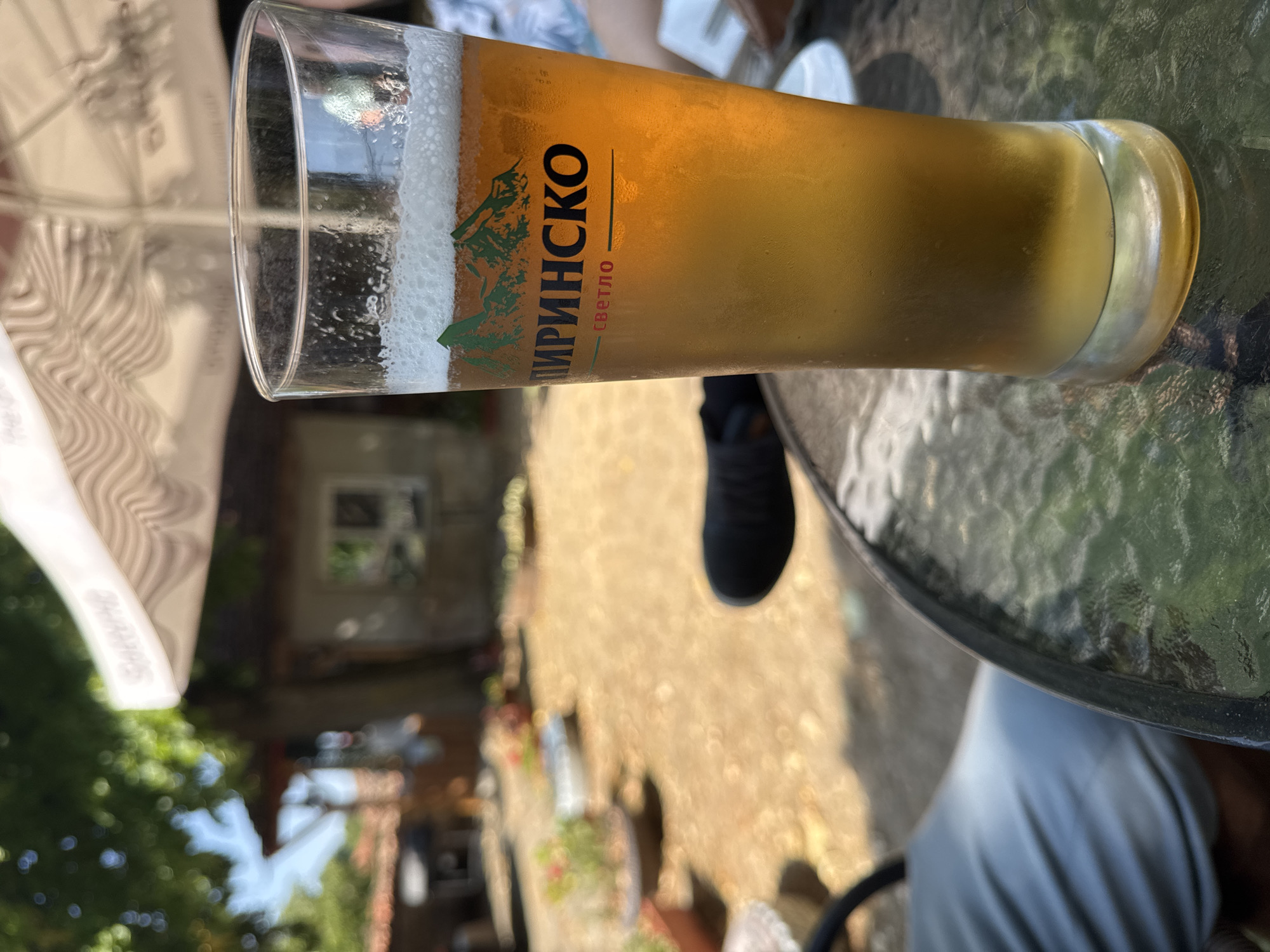 A close-up, eye-level shot features a tall glass of light golden beer with a foamy head, frosted condensation on the glass, and condensation droplets. The glass rests on a textured glass tabletop. In the background, blurred out, is an outdoor setting with a patio or courtyard area. A white umbrella canopy is visible above and behind the beer glass. The area appears to be in Zheravna, Bulgaria, possibly at an outdoor dining establishment. The image captures a relaxed atmosphere on a sunny day.