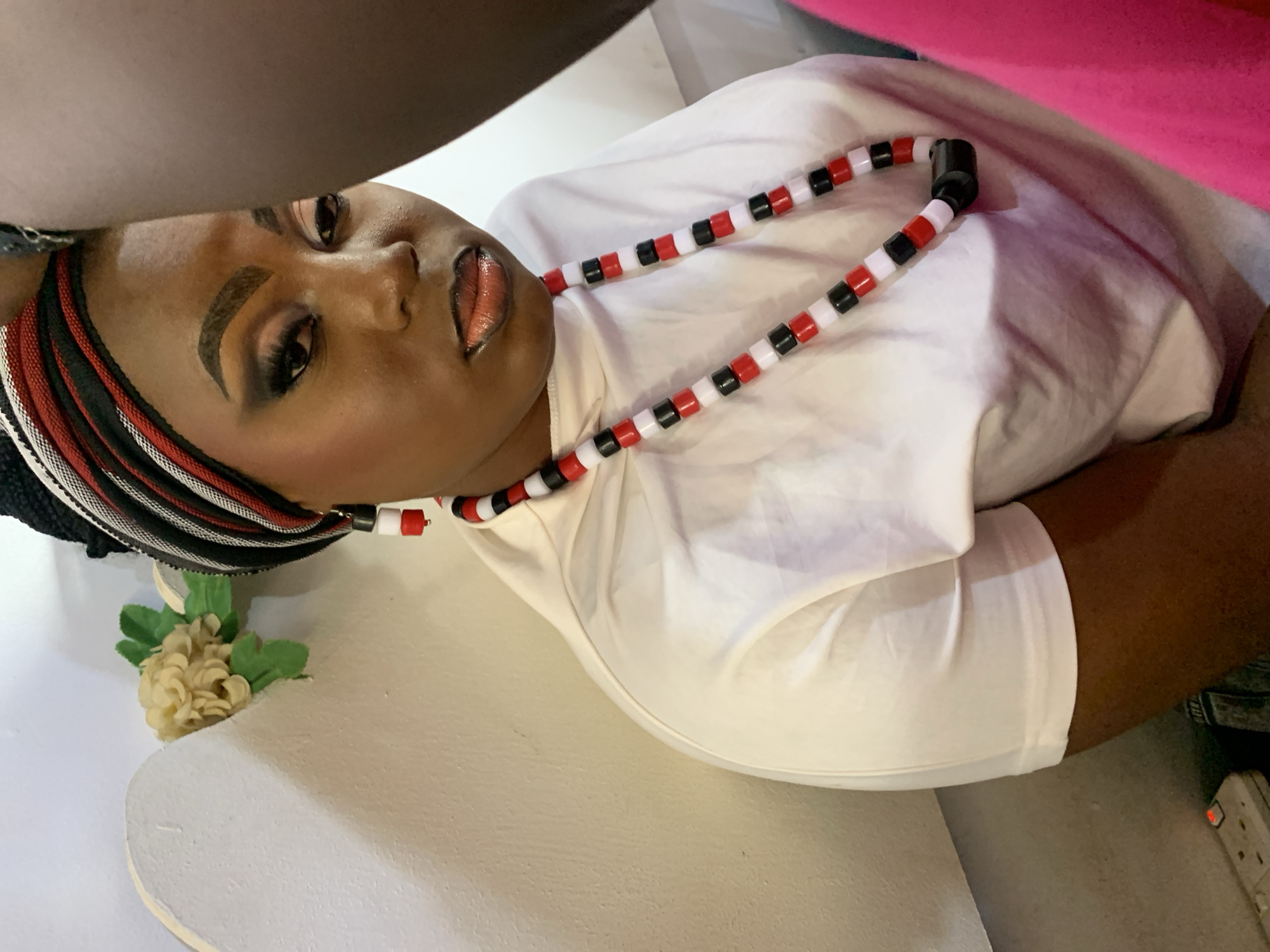 A woman is lying on her side in what appears to be a room. She is wearing a white t-shirt and a beaded necklace with red, white, and black beads. She also has a head wrap with similar colors. The woman is heavily made up, and her lips are parted. In the lower left corner of the image, there is a small bouquet of artificial flowers. The image is taken from a high angle. The location is presumed to be Jos, Nigeria.