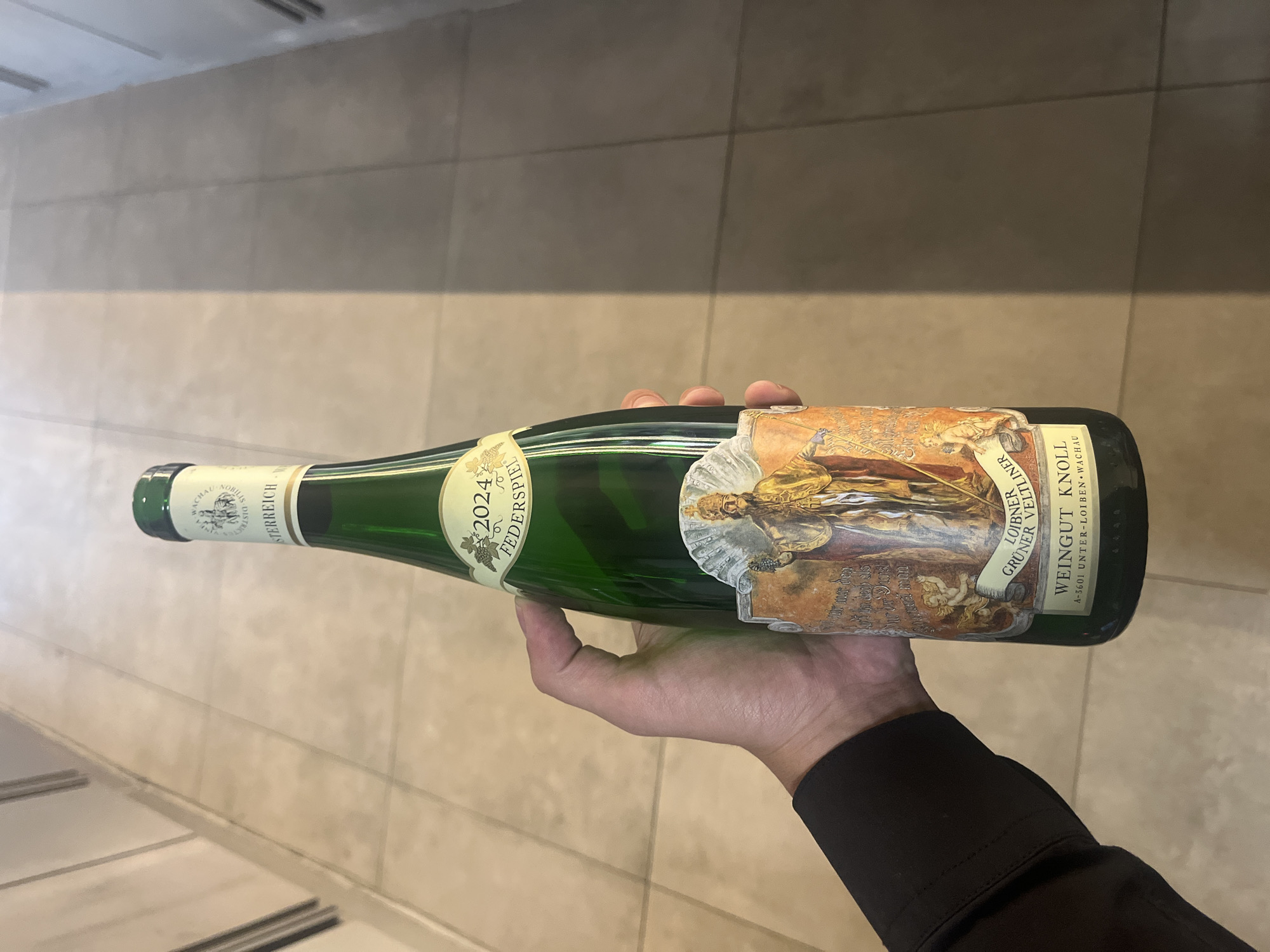 A close-up shot shows a person's hand holding a green glass wine bottle horizontally against an indoor background of light-colored, possibly tiled, floor and walls. The hand is visible from the wrist, which is covered by a dark sleeve. The lighting is bright and even, consistent with an indoor setting during the day.
The wine bottle features an elaborate, multi-part label. The main label is highly decorative, showcasing an intricate, historical-style illustration with figures and architectural elements. Key visible text on the label identifies the wine: "WEINGUT KNOLL" (the winery), "GRÜNER VELTLINER" (the grape variety), "LOIBNER" and "WACHAU" (regions of origin), and "ÖSTERREICH" (Austria). The vintage year "2024" is prominently displayed, along with "FEDERSPIEL," indicating a specific quality level within the Wachau appellation. The winery's address, "A-3601 UNTER-LOIBEN WACHAU," is also printed.
The image captures the presentation of the wine bottle, with its distinct labels clearly visible. The photograph was taken in Sofia, Bulgaria.