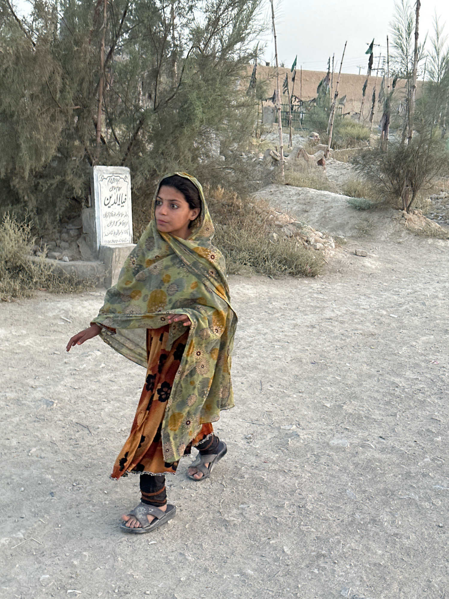 A young girl stands in a dirt area, her body angled to the left and her head turned to the right. She is wearing a green and yellow floral patterned shawl over an orange skirt with a black floral pattern and black leggings. Her feet are clad in grey sandals. To her left, a white stone marker stands upright. In the background, a cluster of thin poles with tattered flags are visible, set against a backdrop of dusty, dry terrain with sparse vegetation and some low-lying structures. The ground is a mixture of sand and gravel.