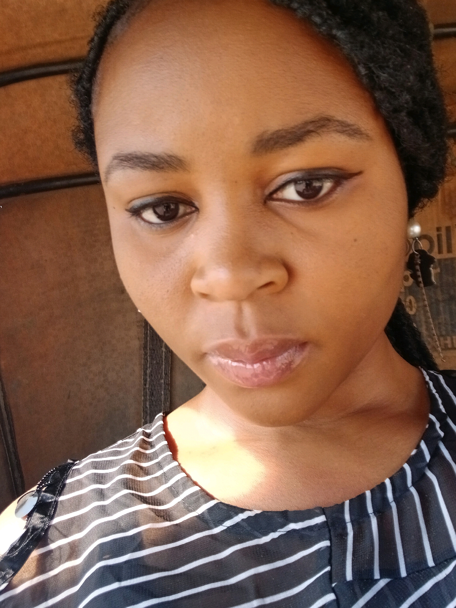 A close-up, slightly angled, selfie-style photograph shows a young Black woman's face and upper torso, indoors. The woman has dark skin, dark eyes, and dark hair braided and pulled back. She is wearing a black and white striped shirt. The background is a blurry, neutral-toned wall with horizontal dark lines. A distinct shadow falls across the woman's right cheek and chest, suggesting artificial lighting or sunlight coming from the right. The image appears to have been taken in an urban environment, potentially within a home or vehicle. No specific landmarks or environmental features definitively place the location in Rayfield, Nigeria, though the style of the background elements is consistent with such an environment.