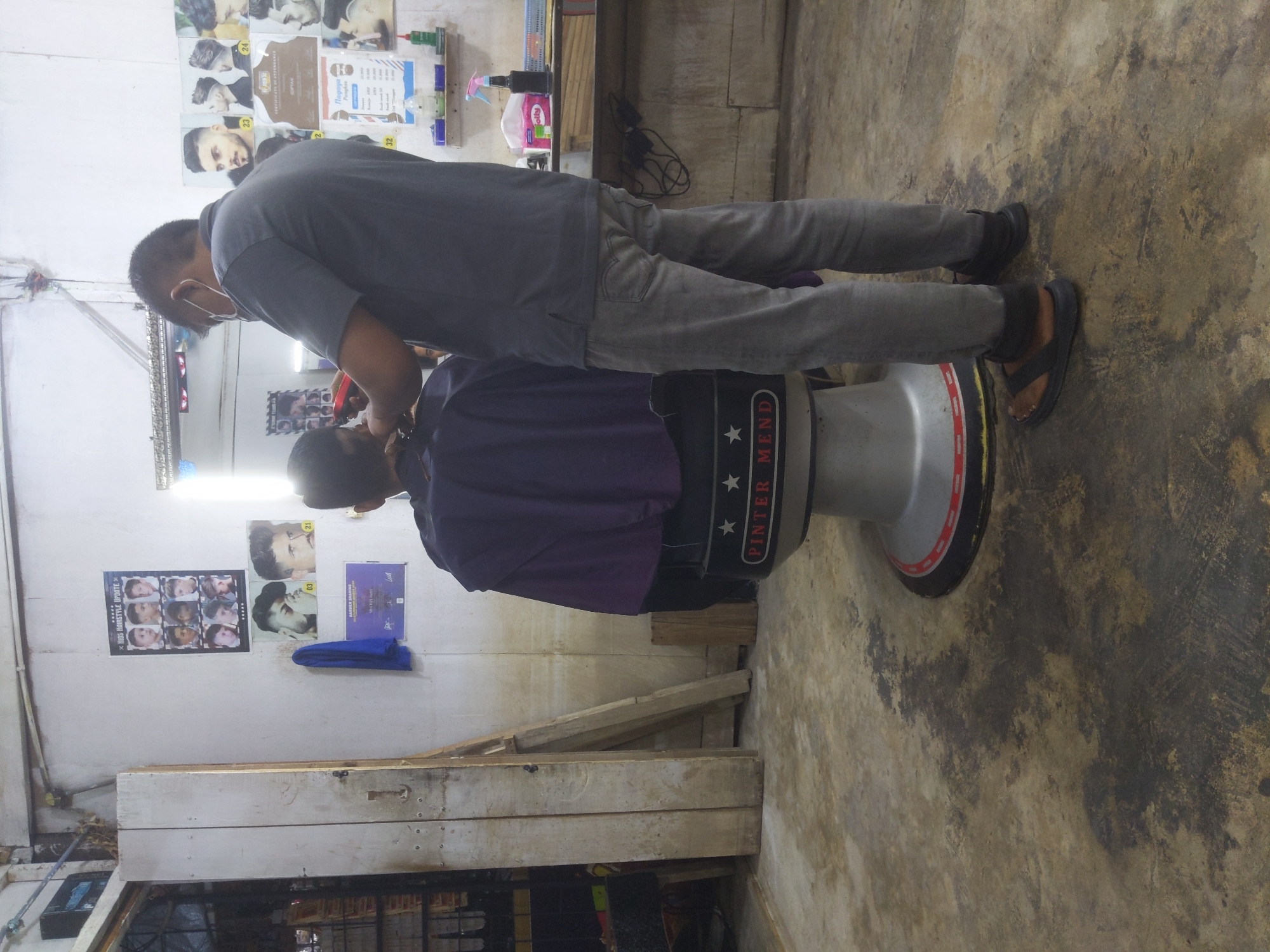 This image depicts the interior of a barbershop in Keudematangpanyang, Indonesia. A male barber, wearing a grey t-shirt, grey pants, a face mask, and sandals, is standing and cutting the hair of a customer. The customer is seated in a barber chair draped with a dark purple cloth. The barber chair has a silver base with red and white accents and branding that reads "PINTER MEND" with three star icons. The walls of the barbershop are decorated with various posters featuring hairstyles and certifications. To the left, there is a wooden door and a wooden structure leaning against the wall. The floor is concrete and appears worn and stained. On a shelf in the background, there are bottles of grooming products, including a spray bottle and tissues.