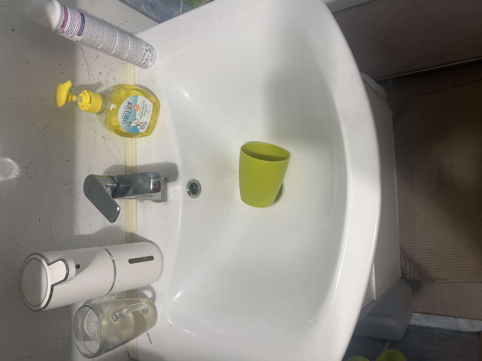 A white ceramic sink is shown in a bathroom, featuring a chrome faucet, a white automatic soap dispenser, a glass filled with water, a bottle of yellow liquid soap, and a green cup. The sink area also includes a white spray can and a brown bath mat. The scene is set in Surgut, Russia, within a residential bathroom.