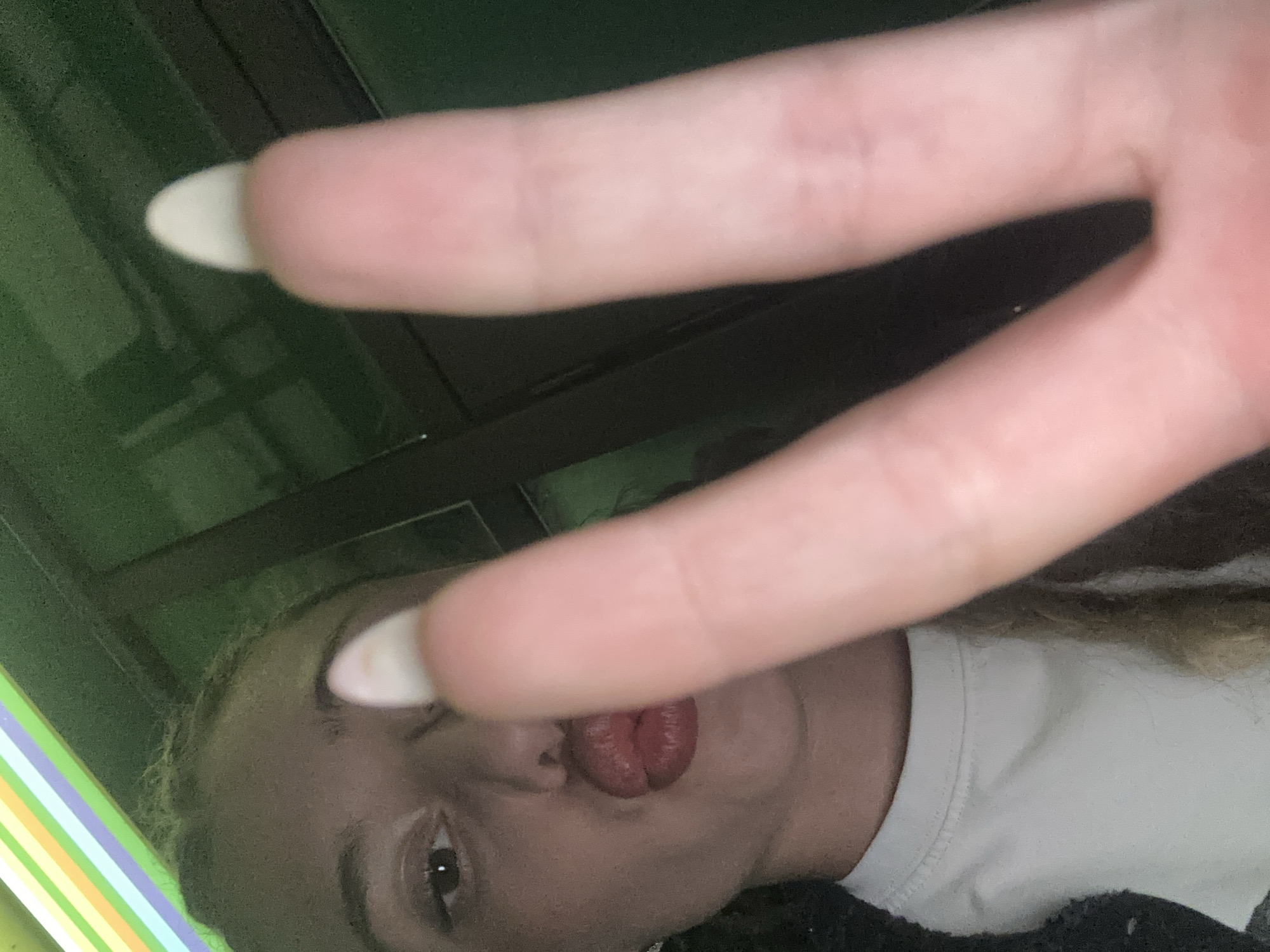 The image is a close-up selfie of a young woman with her lips puckered in a kiss, holding up two fingers in a peace sign gesture. Her right hand, with long, almond-shaped fingernails, is prominently featured in the foreground, obscuring part of her face. She is wearing a white t-shirt. The background appears to be an indoor environment with green walls and a striped element at the top, suggesting it might be a booth or a decorated area. There is no discernible information in the image that would place this scene in Gabrovo, Bulgaria, or any specific city.