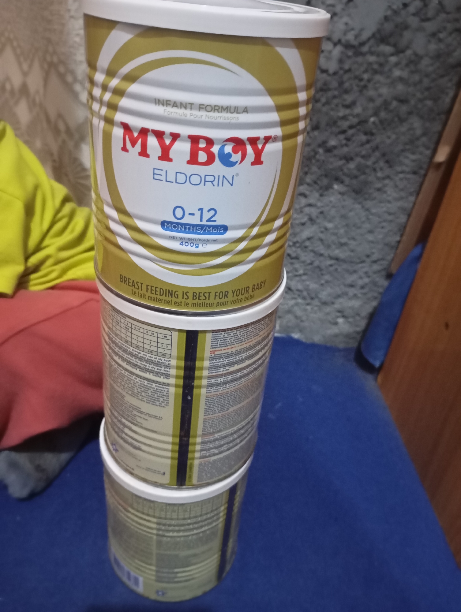 A stack of three cylindrical baby formula cans sits on a dark blue surface, likely a piece of furniture. The top can is predominantly white with gold and red accents, featuring the brand name "MY BOY" in large red letters and "ELDORIN" underneath. Text on this can indicates "INFANT FORMULA," "0-12 MONTHS/Mois," and "400g e." Below this, the text "BREAST FEEDING IS BEST FOR YOUR BABY" is visible.
The second and third cans are partially obscured but appear to have similar branding, with vertical yellow and dark blue stripes. The second can, stacked directly on top of the third, shows nutritional information tables and text in multiple languages. The overall setting appears to be indoors, with a textured gray wall and a hint of a patterned wallpaper in the background on the left. A person wearing a bright yellow hooded garment and a red garment is partially visible on the left side of the image. The lighting suggests an indoor, possibly residential, environment.