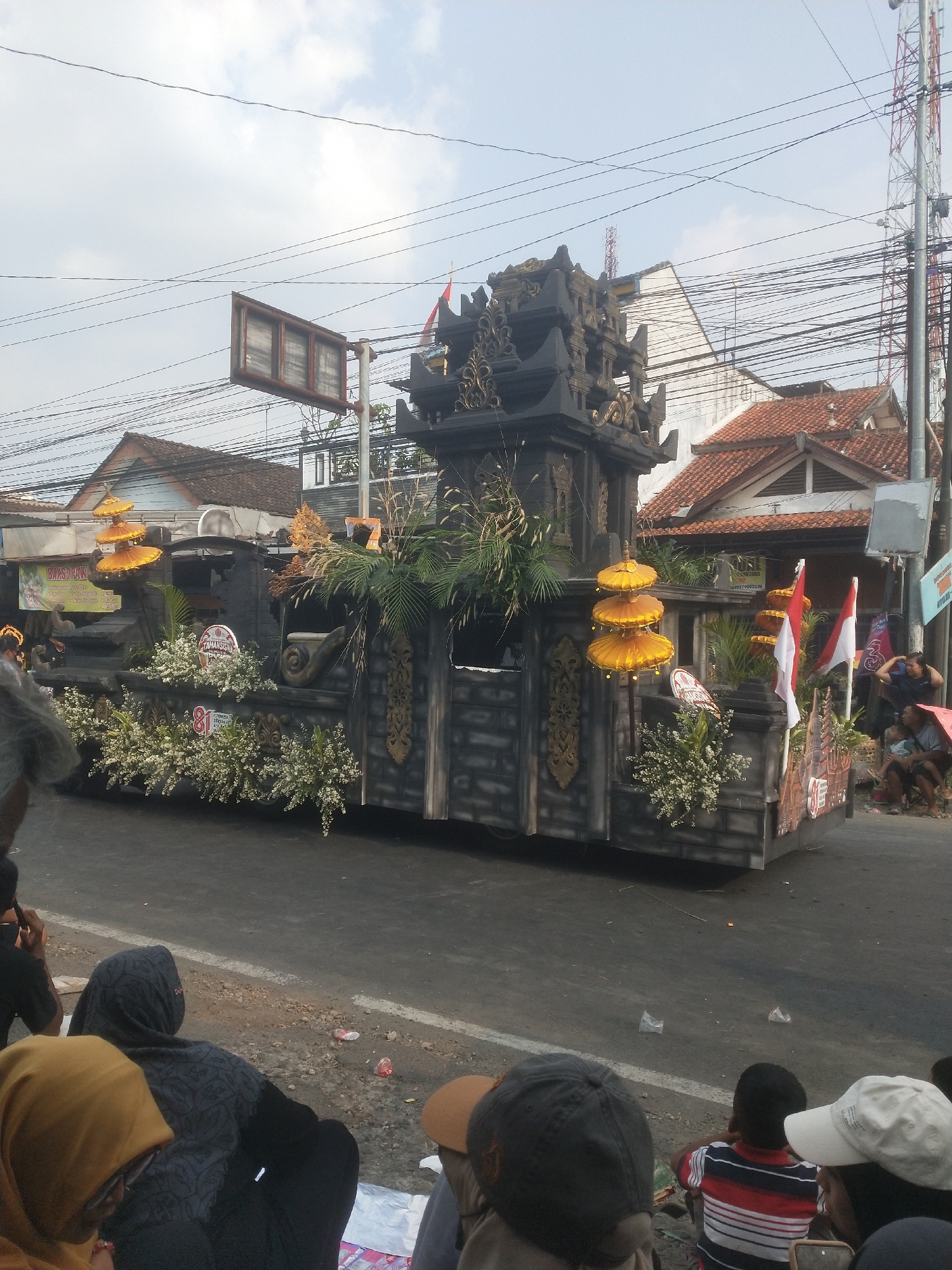 In Boja, Indonesia, a street is lined with spectators watching a parade float. The float is ornately decorated, resembling a dark, tiered temple structure with golden accents and yellow, umbrella-like ornaments. White flowers and green foliage adorn the float's base. Two Indonesian flags, red and white, are prominently displayed on the right side of the float.
A crowd of people is seated on the side of the road, facing the float. They are dressed in casual clothing, and some are wearing head coverings. A few individuals are standing further back, observing the procession. The street itself appears to be paved, with a white line marking the edge. Buildings with red-tiled roofs and various power lines are visible in the background, indicating an urban or semi-urban setting. The sky is partly cloudy. A large traffic signal pole with multiple signs is also visible on the right side of the image.