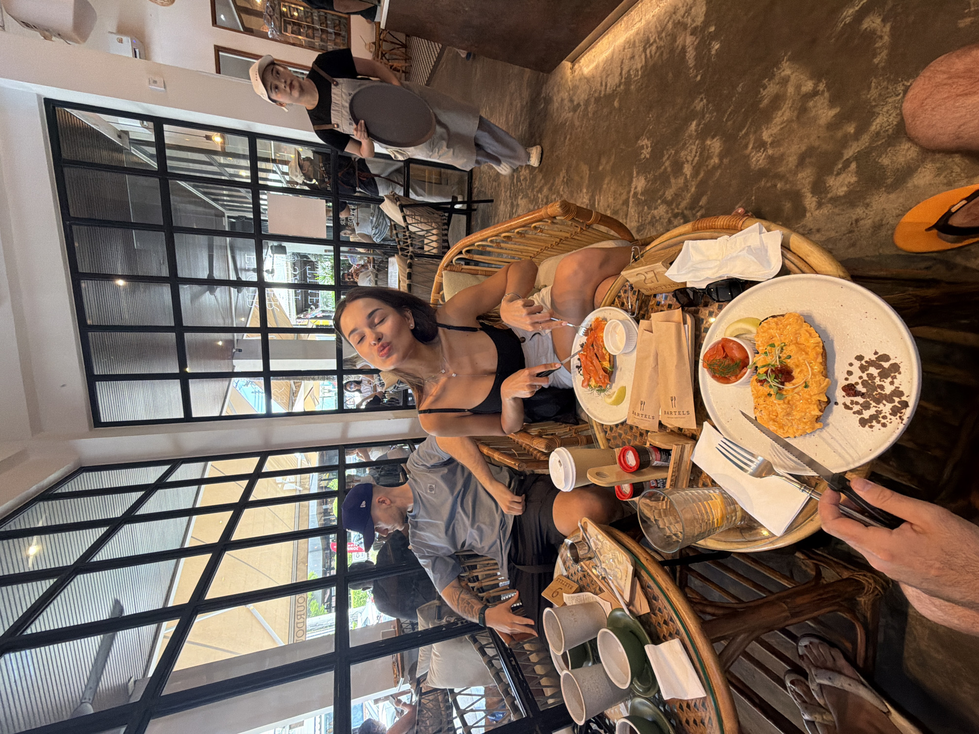 The image shows an indoor dining scene at what appears to be a cafe in Choeng Thale, Thailand, identifiable by the "Bartels" branding on the food packaging. A young woman is seated at a table with food, making a kissing face towards the camera. The setting includes wicker furniture and tables. A man in a blue cap is seated across from her, and another individual is partially visible on the right side of the frame, holding cutlery near a plate of food. A waitress in a white cap and apron stands in the background, holding a tray. The cafe has a large window structure, providing a view of the exterior. The overall lighting suggests a daytime setting. The scene implies a casual dining experience.