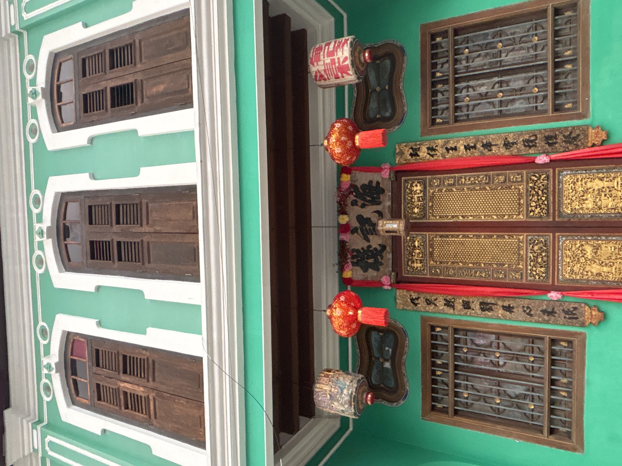 The image shows the facade of a traditional building in George Town, Malaysia. The building is painted a vibrant turquoise green with white trim. On the upper level, there are three arched wooden windows with shutters. Below these windows, a wooden overhang supports decorative elements. On the lower level, there is a large, ornate wooden door, gilded with intricate carvings and patterns. Flanking the door are two windows with metal grilles. Red silk curtains hang on either side of the door, and two decorative lanterns are suspended from above. One lantern is red and round with tassels, while the other is cylindrical and white with red Chinese characters. A wooden plaque with Chinese calligraphy hangs vertically next to the door. The overall architectural style suggests a Chinese shophouse, common in George Town's historic districts.