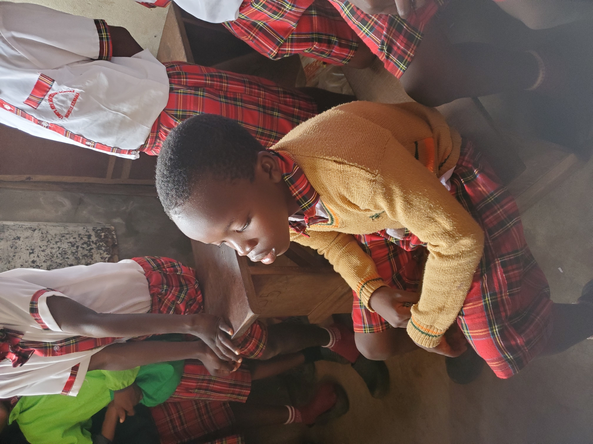 The image captures an indoor scene, likely a classroom in Ukwu-Abwa, Nigeria, filled with several children. The primary focus is on a child in the foreground, positioned centrally, wearing a mustard yellow knitted sweater over a uniform. This child is looking downwards, appearing focused or engaged with something on the wooden desk before them.
Other children, partially visible, are seated around similar wooden desks. They are dressed in a distinctive school uniform consisting of white shirts with red and plaid trim on the sleeves and collar, paired with red and black plaid skirts or kilts. Some children are also wearing red socks with white stripes and dark shoes or sandals. On one of the white shirts, text is visible, identifying the institution as "LORD'S MERCY SCHOOLS" with "MWELU EZEMKA" written below it.
The setting appears modest, with a concrete floor and simple wooden furniture. The lighting suggests an indoor environment, possibly during the daytime. The children, exhibiting dark skin tones, are depicted in various states of attention, contributing to the ambiance of a busy school day.