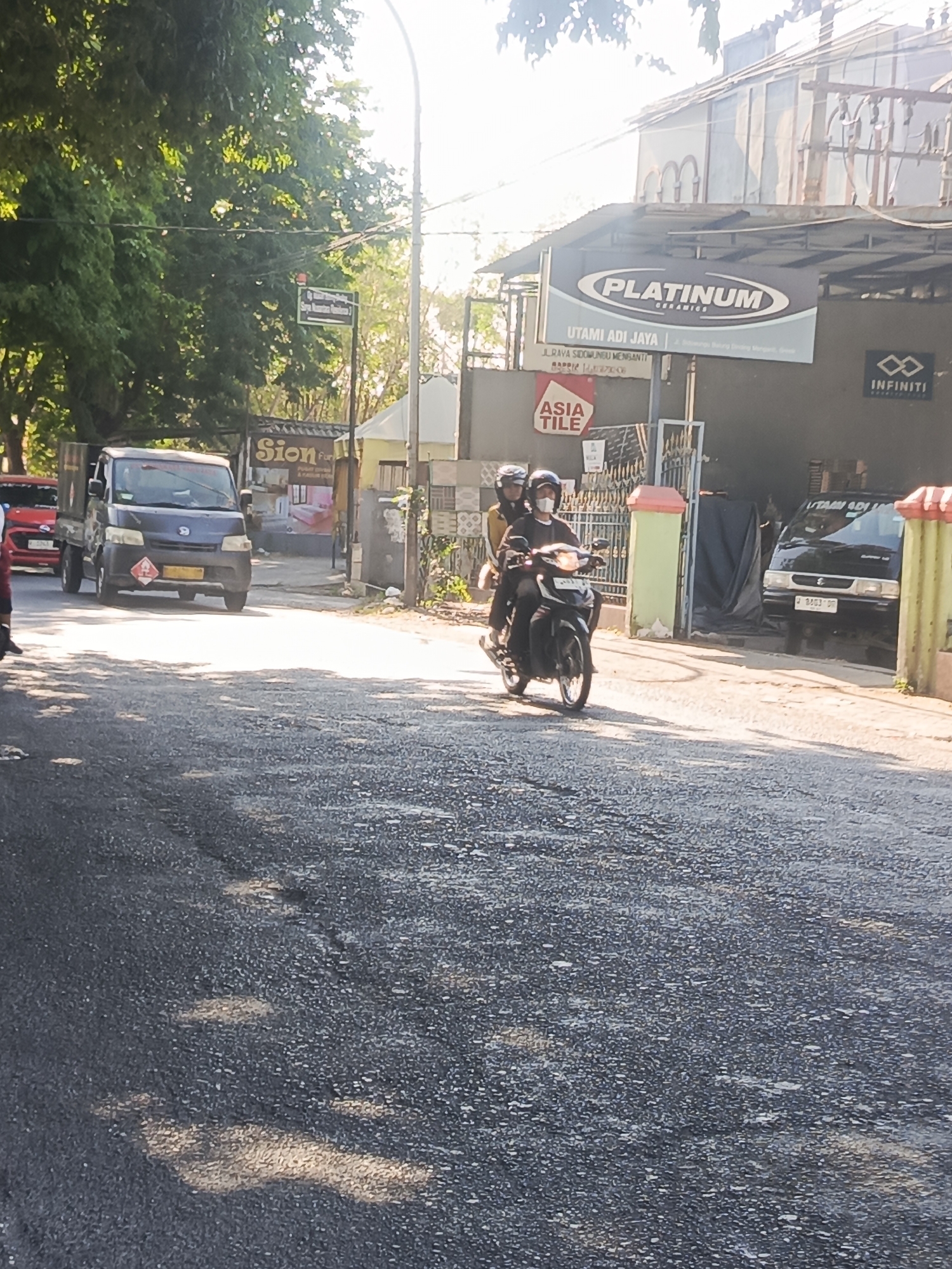A street scene is depicted in Sidowungu, Indonesia, on a sunny day. In the foreground, a gray asphalt road shows signs of wear and tear, with patches of lighter gray and scattered debris. Sunlight casts dappled shadows across the road, suggesting trees line the area.
Several vehicles are visible. A dark blue utility truck is parked on the left side of the road, displaying a yellow hazard placard on its front bumper. To its left, the rear of a red vehicle is partially visible. Further down the road, a black utility truck is parked, its license plate reading "B 8363 OP".
A motorcycle carrying two people, both wearing helmets, is moving away from the camera. The rider is wearing a dark long-sleeved shirt, and the passenger is wearing a yellow jacket. Both appear to be wearing face masks.
Various commercial signs are present. A prominent sign for "PLATINUM CERAMICS" by "UTAMI ADI JAYA" is mounted above a building. Another sign reads "ASIA TILE". A sign for "Sion Fur" is also visible. The building facade behind the "PLATINUM" sign has arched architectural elements. To the right, a sign for "INFINITI" is displayed.
The environment is characterized by a street with trees and buildings lining the sides. Power lines are strung overhead. The overall impression is a commercial street in an Indonesian town.