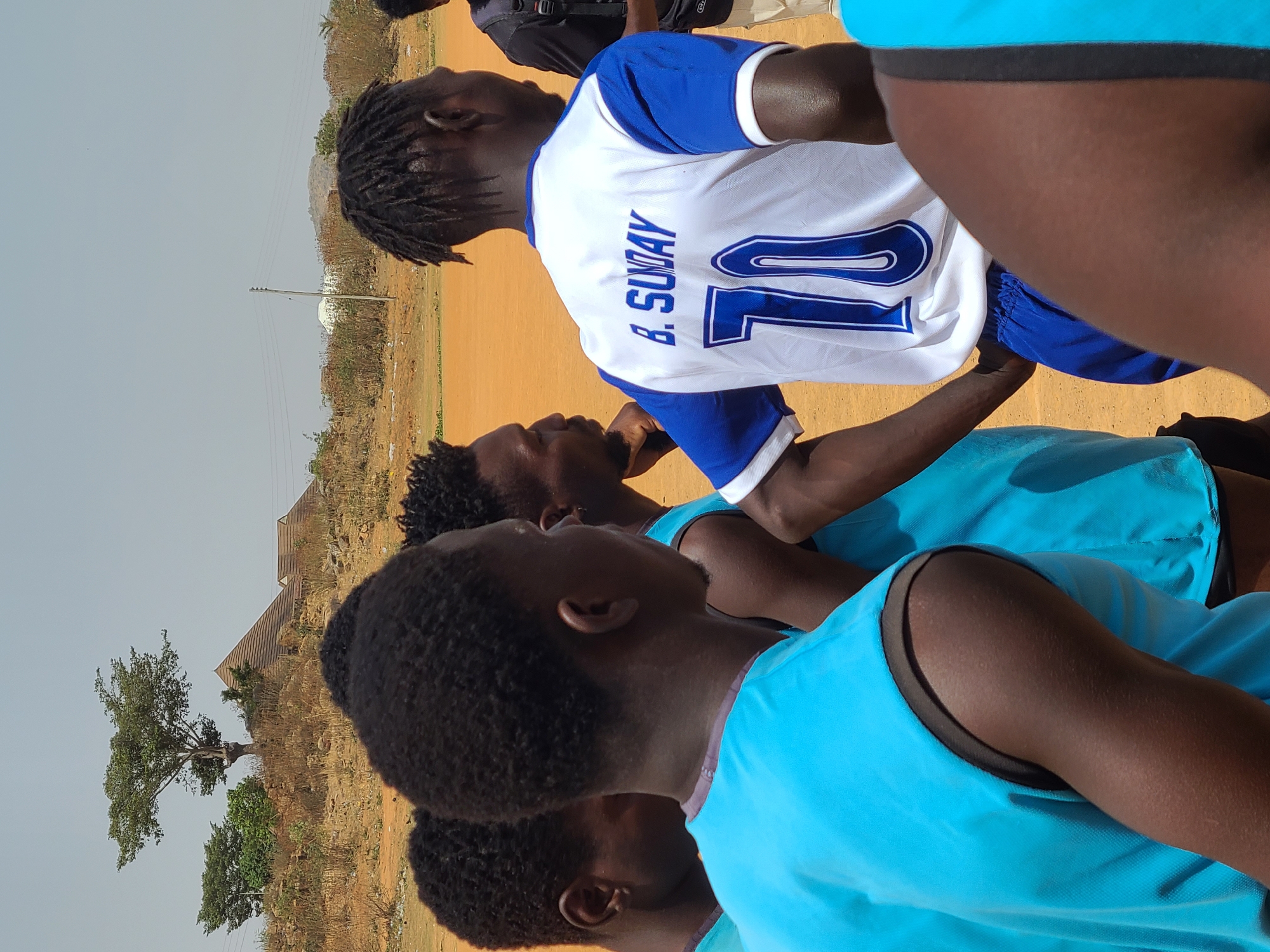 The image captures a group of dark-skinned individuals, likely athletes, gathered on a sunny day in an arid, open field in Barikin Jirga Summa, Nigeria.
In the foreground, several people are partially visible, wearing sports attire. The most prominent figure, seen from behind, wears a white and blue short-sleeved jersey with the name "B. SUNDAY" and the number "10" printed on the back in blue. This individual has dark dreadlocks. Other individuals in the foreground and midground are wearing bright turquoise/light blue sleeveless vests, suggesting they are part of a team or group engaged in a sports activity or training. They appear to be looking towards the person in the white and blue jersey or slightly away.
The setting is an unpaved, reddish-brown dirt field, characteristic of a dry, outdoor environment. The sky is clear and light blue, indicating bright daylight and good weather. In the background, beyond the field, there is sparse, dry vegetation, including bushes and a few trees with green foliage. Distant structures, possibly small buildings or houses, can be seen, along with a power pole. The overall impression is that of a sports practice or casual game taking place in a warm, dry climate.