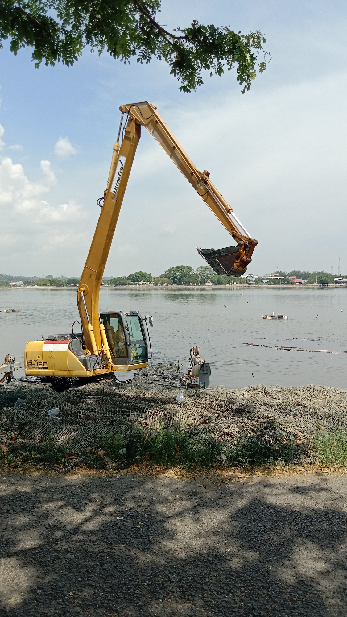 A yellow UltraTre SH130 excavator sits on a pile of fishing nets at the edge of a body of water, possibly a lake or river, in Lhokseumawe, Indonesia. The excavator's arm is extended over the water, with its bucket partially submerged. There is trash floating on the water's surface. The shoreline across the water features trees and buildings. A tree overhangs the excavator from the top of the frame. The ground beneath the fishing nets is paved and covered in shadows. The sky is partly cloudy.