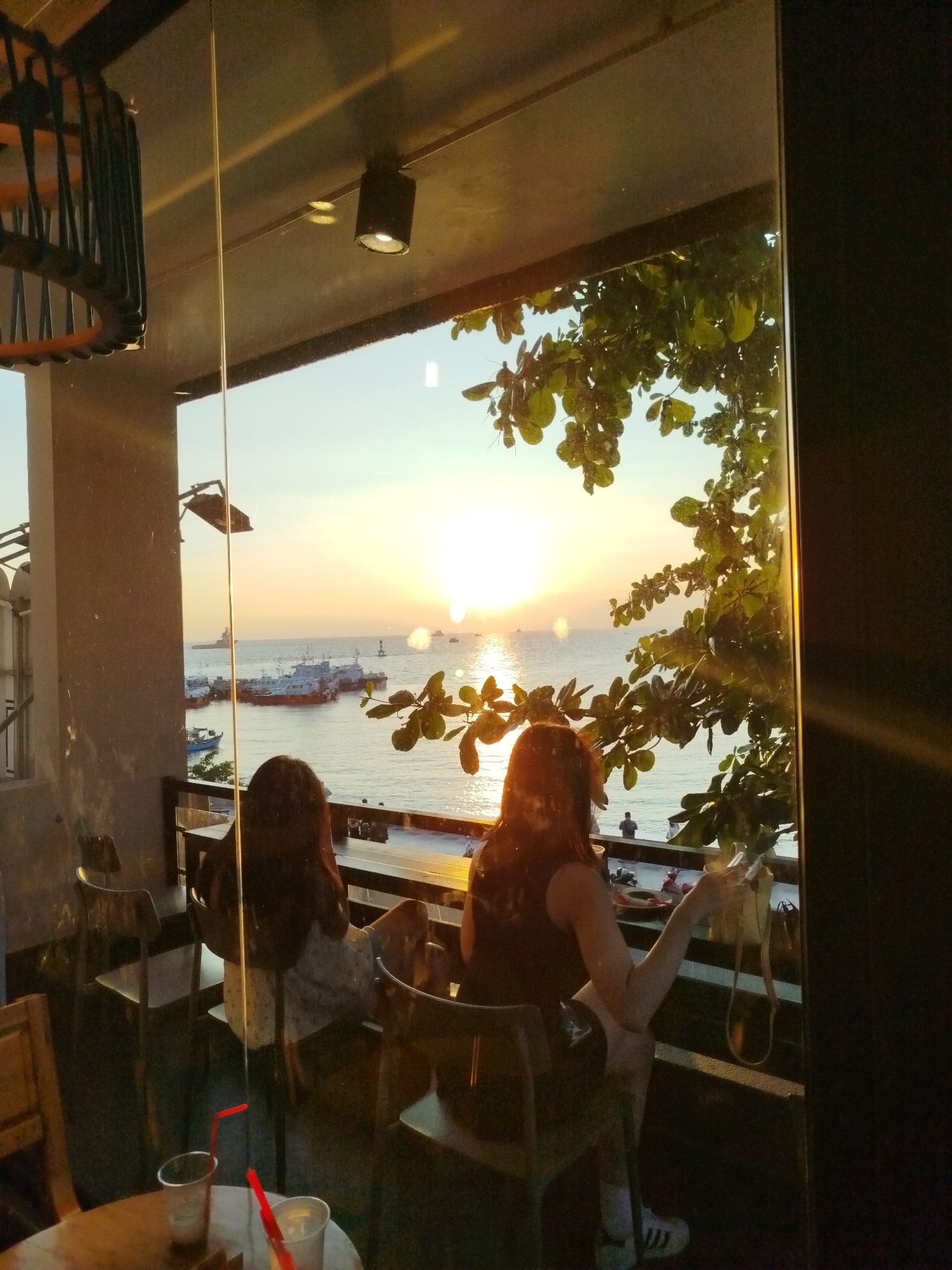 Here is a description of the image:
Two women are seated at a table near a window overlooking the sea and the setting sun. The scene appears to be from a building with a balcony or a large window. Several boats are visible in the sea. There is a tree branch with green leaves partially obstructing the view from the window. The reflections on the glass suggest that the location may be in Ho Chi Minh City, Vietnam. Two drinks with red straws are on the table. One woman is holding a small bag.