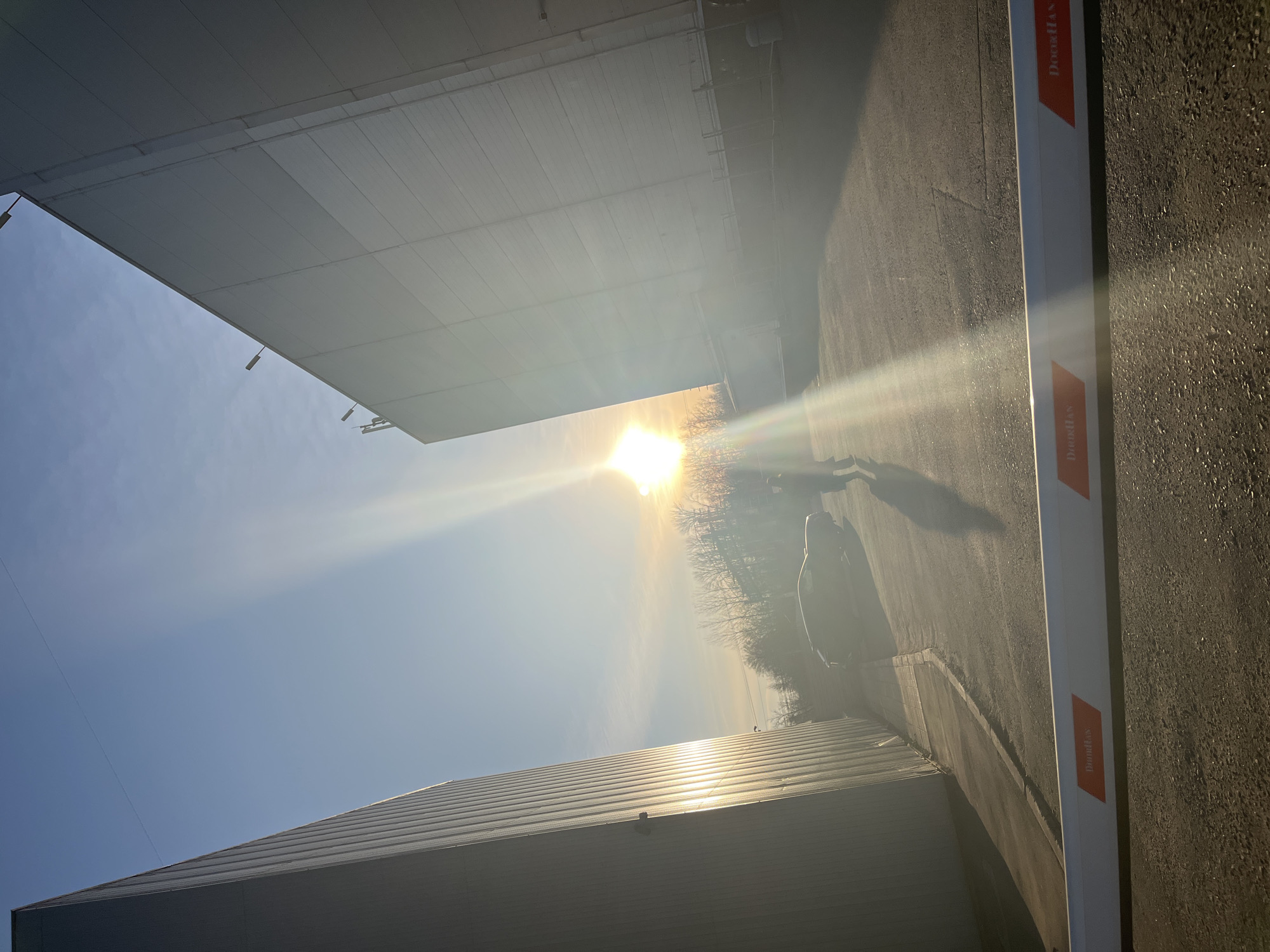 The photograph, taken at ground level, presents a distorted perspective due to an upward angle. The sun, a prominent feature, is positioned between two large, rectangular structures, causing lens flare. The structure on the left, under a blue sky, appears to be a large building with a flat roof, possibly an industrial or commercial facility. On the right, a white barrier arm, labeled with an orange "DormaKaba" logo, is visible. The asphalt ground, showing shadows of people, vehicles, and trees, leads to a patch of sparse trees and bushes under the sun. The overall environment seems industrial or suburban. While no specific details conclusively identify the location as Klin, Russia, the architectural elements and overall environment are consistent with urban and industrial areas.