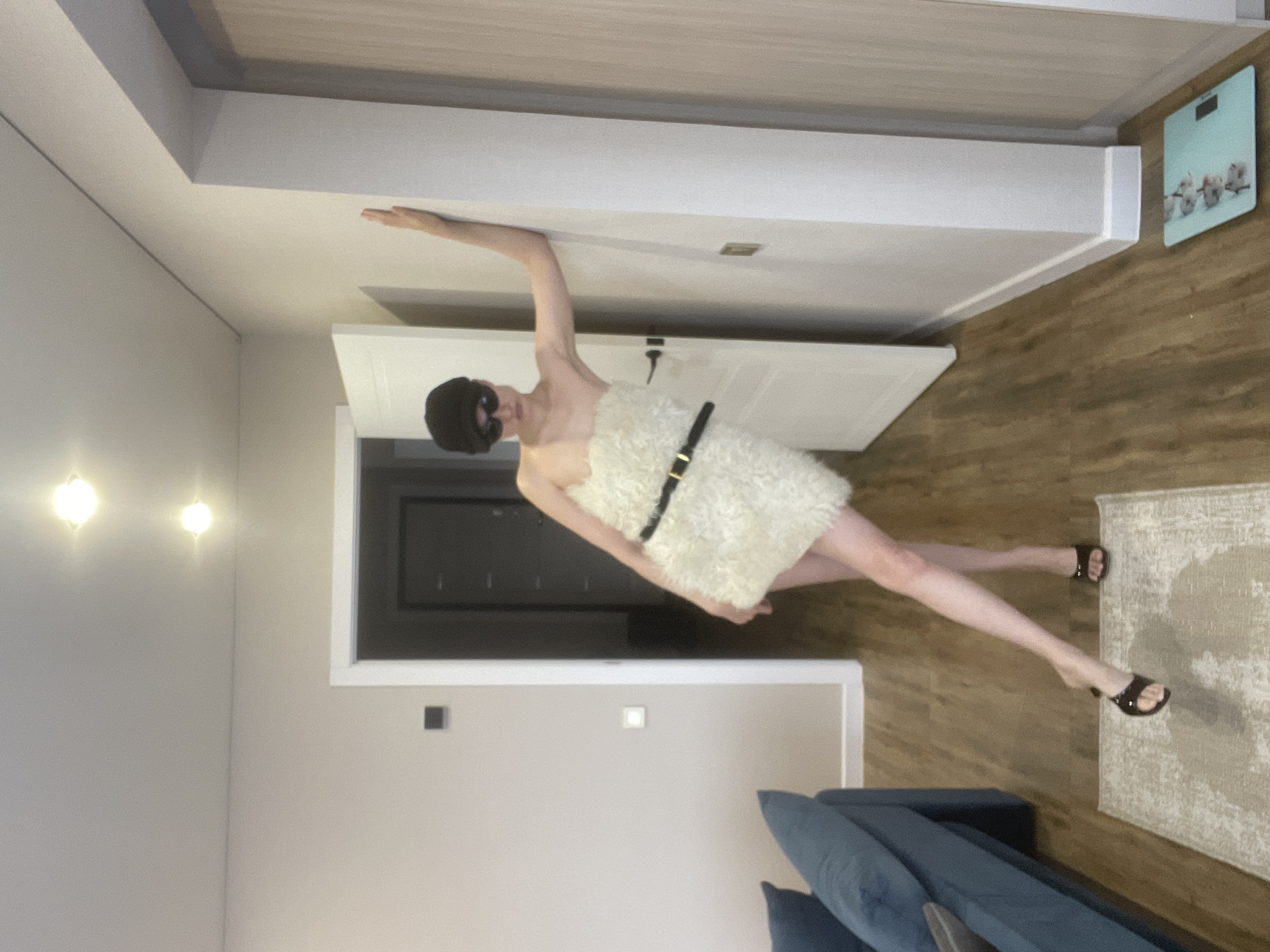 A person wearing a white, fur-like garment secured with a black belt and a black eye mask is leaning against a white wall. Their right arm is extended towards the wall near the ceiling, and their legs are positioned such that one foot is on a rug and the other is near a doorway. The scene appears to be inside a home with white walls and a door visible in the background. A blue couch is also visible in the lower part of the frame. There is also what looks like a digital scale on the top right. Podshivalovo, Russia is not discernible from the image.