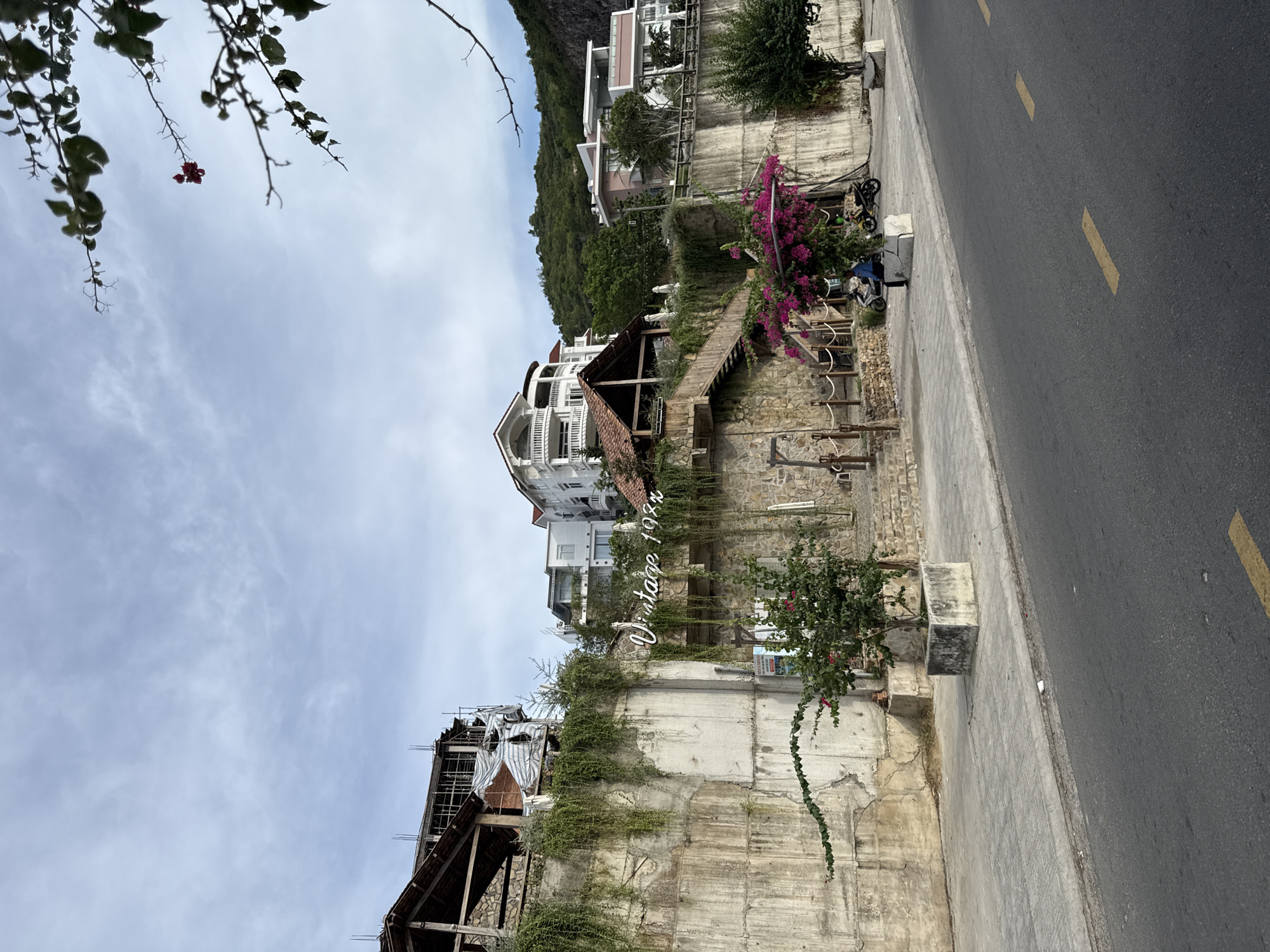 The image shows a street scene in Nha Trang, Vietnam, featuring a multi-story building under construction, a concrete retaining wall covered in hanging plants, and a building with a "Vintage 1988" sign. A wooden staircase leads up the retaining wall to a covered structure. In the foreground, there is a sidewalk and a road with yellow markings. A person is seated on a concrete planter near two motorcycles. Pink bougainvillea flowers are visible. The sky is cloudy. The buildings appear to be situated on a hillside, overlooking the road.