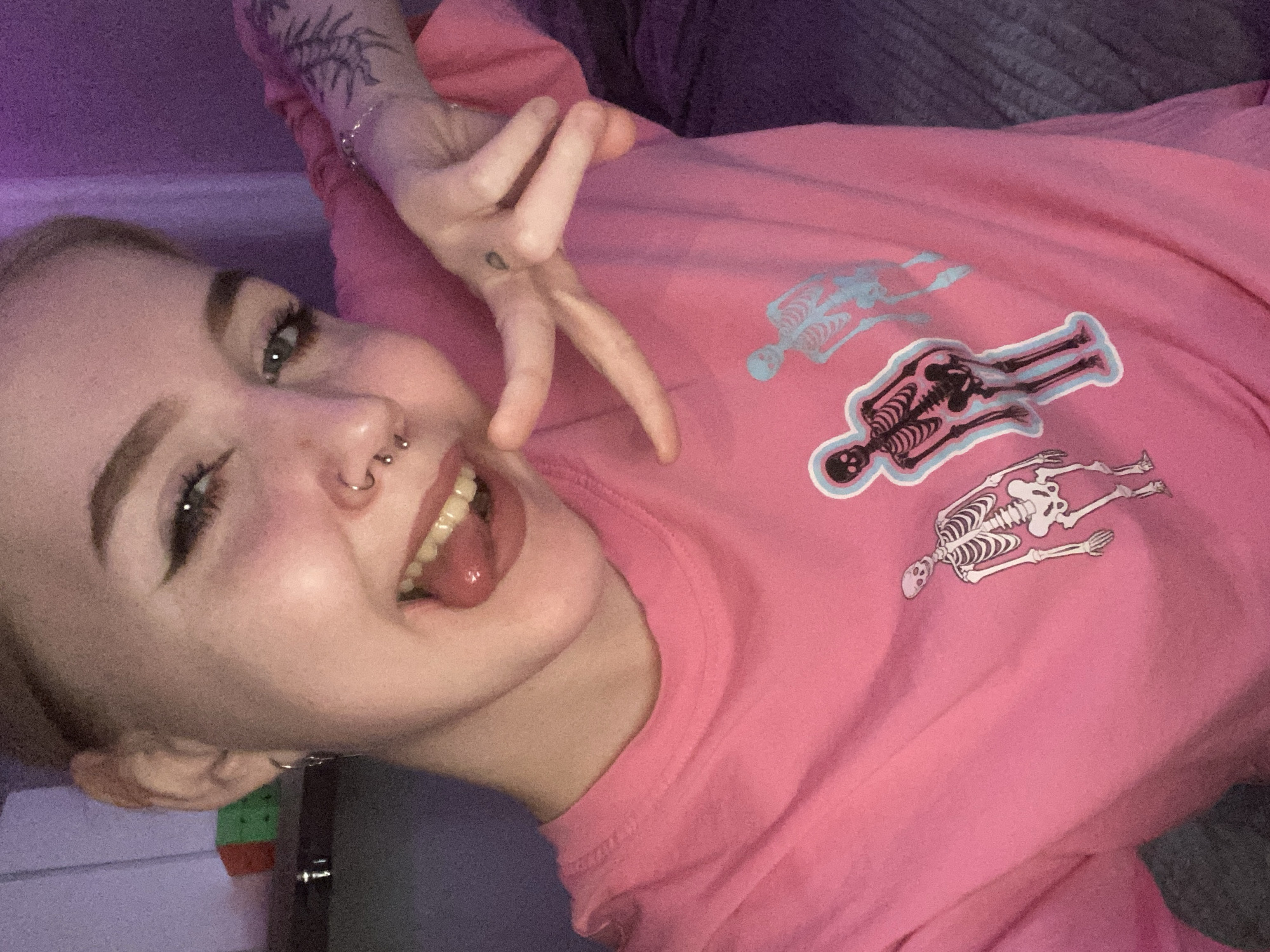 A young woman is posing for a selfie. She is lying down, wearing a pink long-sleeved shirt with three cartoon skeleton decals in different colors on the chest. She has blonde hair, visible nose piercings, and appears to have tattoos on her left wrist and forearm. She is sticking her tongue out and making a hand gesture. The background includes a purple wall and part of a grey textured surface. The location is unknown.