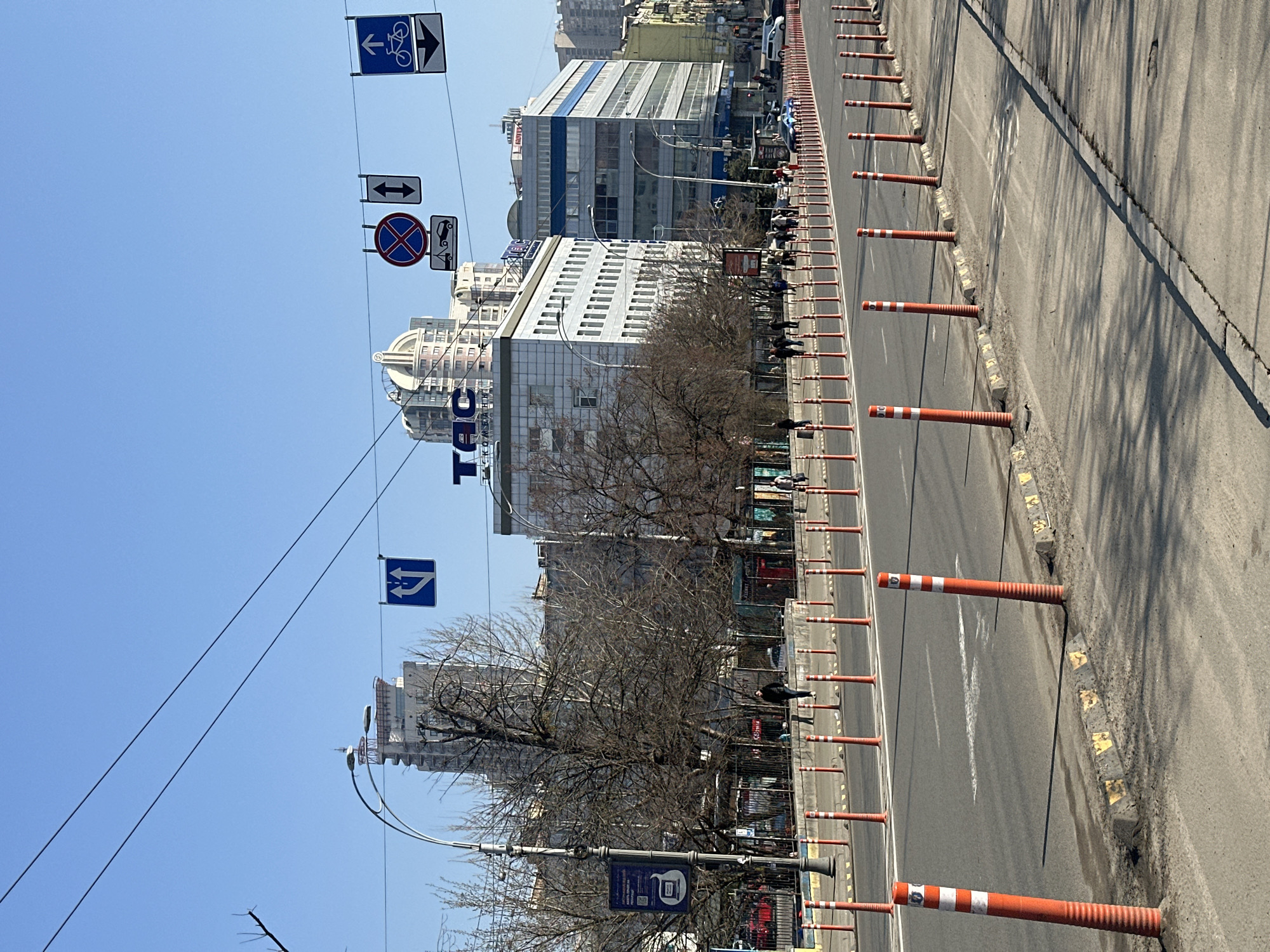 This image depicts a street scene on Velyka Vasylkivska Street, Kyiv, Ukraine, facing generally northwest. The weather is clear and sunny, indicated by a bright blue sky and distinct shadows.
The foreground features a multi-lane asphalt road with white lane markings. The road's median is delineated by a double row of orange and white striped flexible bollards anchored to the pavement, extending into the background. A concrete sidewalk borders the right side of the road, separated by a row of these bollards and small, yellow-and-black painted concrete curbstones. Another sidewalk runs parallel on the left, separated from the road by a similar row of bollards.
On the left side of the street, leafless deciduous trees with dark, gnarled branches are visible behind a black metal fence, suggesting late winter or early spring. A tall, black, ornate street light post with multiple curved arms and fixtures is positioned on the left, partially obscuring the trees and a large digital advertising board with a QR code.
In the mid-ground, on the left side, stands a multi-story building with a light-colored facade and numerous windows, topped by a prominent blue sign displaying "TAC КОМБАНК" (TAC COMBANK). Behind and slightly to the right of this building, a taller, more distant high-rise features a distinctive white, arched, domed roof structure with a finial. To the right of the TAC building, another multi-story commercial building with a grey facade and a horizontal blue stripe near its top is visible. Further down the street, additional buildings of varying architectural styles line the road.
Pedestrian traffic is observed on the sidewalks in the mid-ground and background, with individuals walking in both directions. One person in a dark jacket is clearly visible walking on the left sidewalk. Vehicular traffic is present in the distance on the right side of the road, including a dark blue sedan and a white van.
Overhead wires suspend several rectangular street signs across the intersection:
* A blue square sign with two vertical white arrows indicating opposing directions.
* A circular "no stopping" sign (red cross on blue background) with a supplementary white rectangular sign depicting a tow truck, indicating vehicle removal.
* A blue square sign with an upward-pointing white bicycle symbol and a small downward-pointing arrow.
* A blue square sign with a single downward-pointing white arrow.
