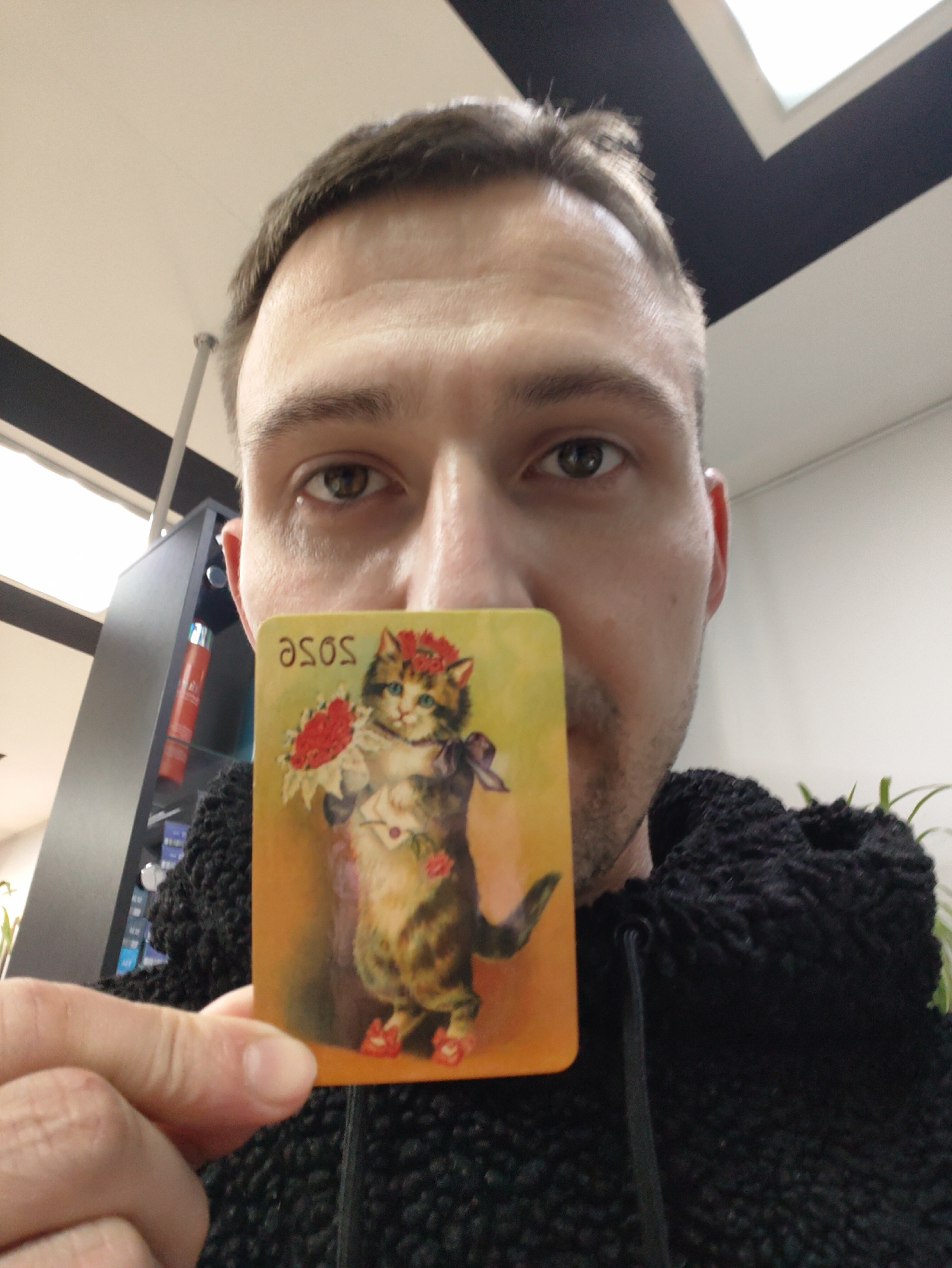 A man is holding a card in front of his face. The card depicts a painting of a cat wearing a floral crown, holding flowers, and wearing red shoes. The card also has the word "asos" printed on it. The man is wearing a black, textured hoodie. The background includes a black cabinet with products on the shelves, a white wall, and a skylight. The location is not specified.