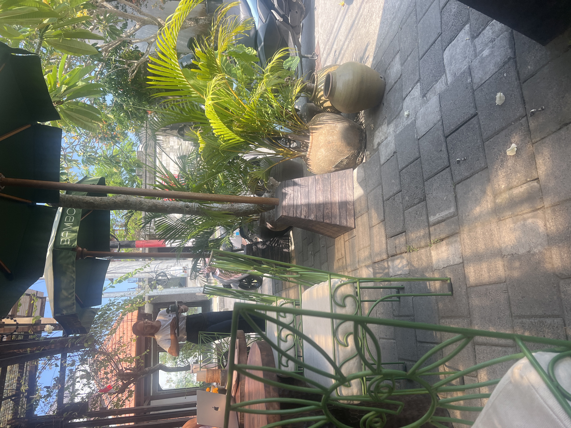 The image depicts an outdoor seating area at a cafe in Mengwi, Indonesia. A woman in a white t-shirt and black apron is serving a tray with what appears to be drinks, indicating she is a server. She is standing near a wooden table where a laptop with an Apple logo is visible. Green metal chairs with white cushions are arranged around the table. In the background, lush tropical plants, including palm trees in large ceramic pots, are present. A motorcycle is parked in the background, suggesting a street or pathway nearby. The ground is paved with gray interlocking stones. Sunlight casts shadows across the scene, indicating it is daytime. The cafe appears to have a relaxed, outdoor ambiance, typical of a tropical location.