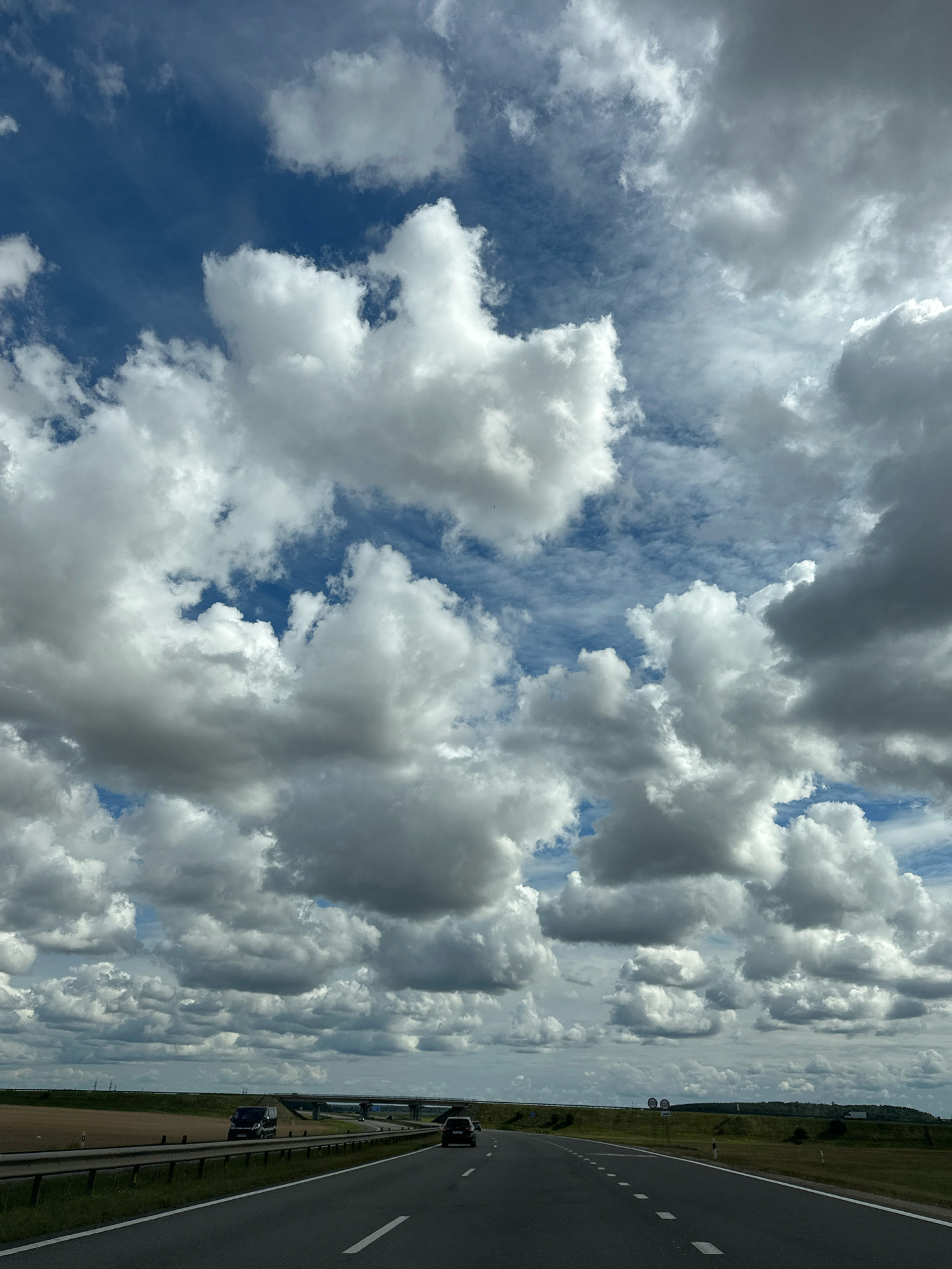 The image shows a view from a moving vehicle on a highway in the direction of travel. The sky is filled with large, fluffy white and grey clouds against a blue sky. The highway has two lanes in the direction of travel, with a white line marking the center and solid white lines on the right edge. There are guardrails on the right side of the road. In the distance, there are some vehicles on the highway and a bridge. The landscape to the right of the highway consists of green grass and some trees. To the left of the highway, there appears to be a field. The location is identified as Blonski Selski Sovet, Belarus.
