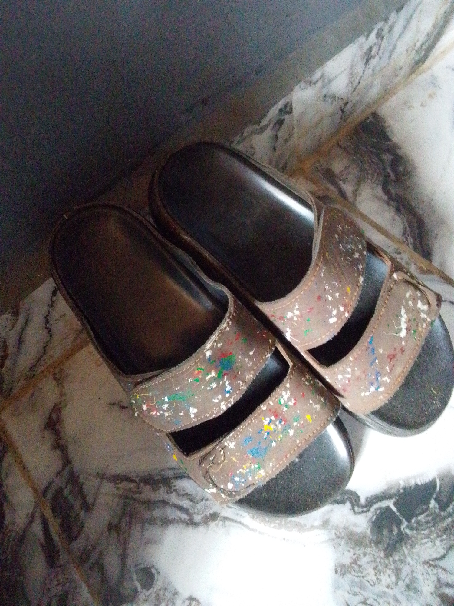 Two open-toed brown sandals with a paint-splatter design are lying on a marble floor. The sandals have two straps across the foot, and the soles are black. The background is dark and out of focus, suggesting the image was taken indoors, possibly in a corner where a wall meets the floor.