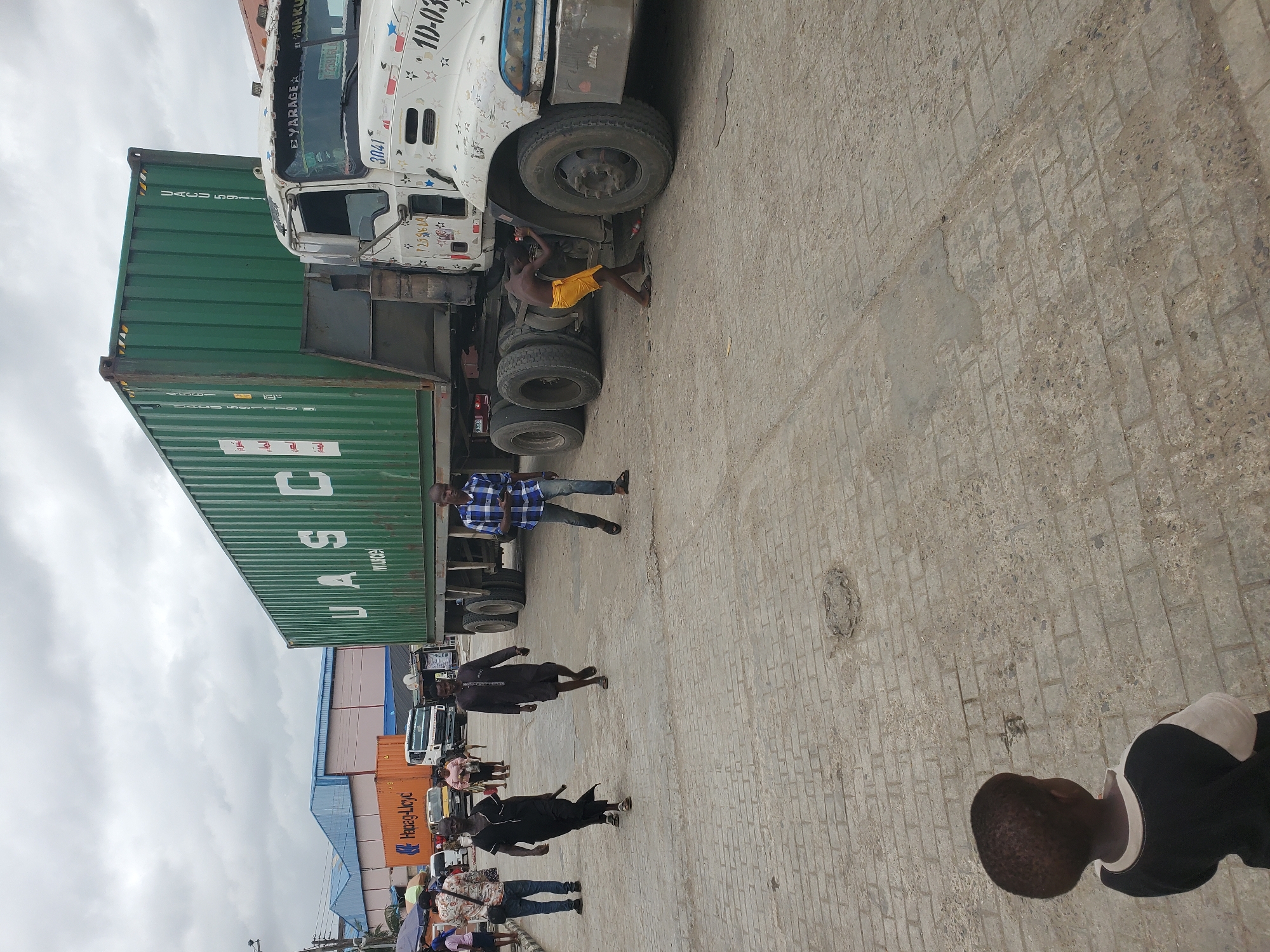 The image depicts a busy outdoor scene in Lagos, Nigeria, likely a port or industrial area. A large green shipping container mounted on a white truck is the primary focus. A man in a yellow cloth appears to be working on the underside of the truck near the front tires. Several other individuals are walking past the truck. These people are dressed in casual attire.
The environment includes a paved area, possibly a street or loading zone, with other stacked shipping containers in the background. The sky is overcast. Text on the container includes "UASC," "WUICE," and "UACU 59," while "Hapag-Lloyd" is visible on a nearby container. The truck's license plate information includes "NA KU," "725916 LA" and "10-03". The truck also has some visible writing on the front cab that is labeled "YARAGE" and "3041".