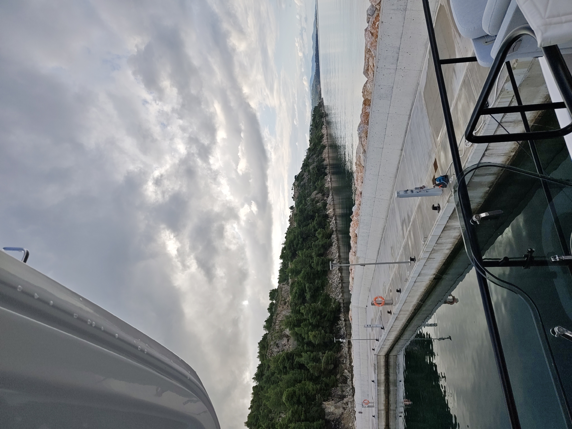 This is a high-angle, wide shot of a modern marina in Sithonia Municipal Unit, Greece, under a cloudy sky. The foreground features the side of a white yacht, angled from the upper left of the frame. Below the yacht, the dark, reflective water of the marina is visible. A concrete pier runs horizontally across the mid-ground, lined with white concrete barriers and topped with orange life preservers and bollards. The pier extends into the distance along a rocky, tree-covered coastline. The sea stretches to the horizon, meeting a hazy, mountainous landscape under a dramatic sky filled with grey and white clouds.