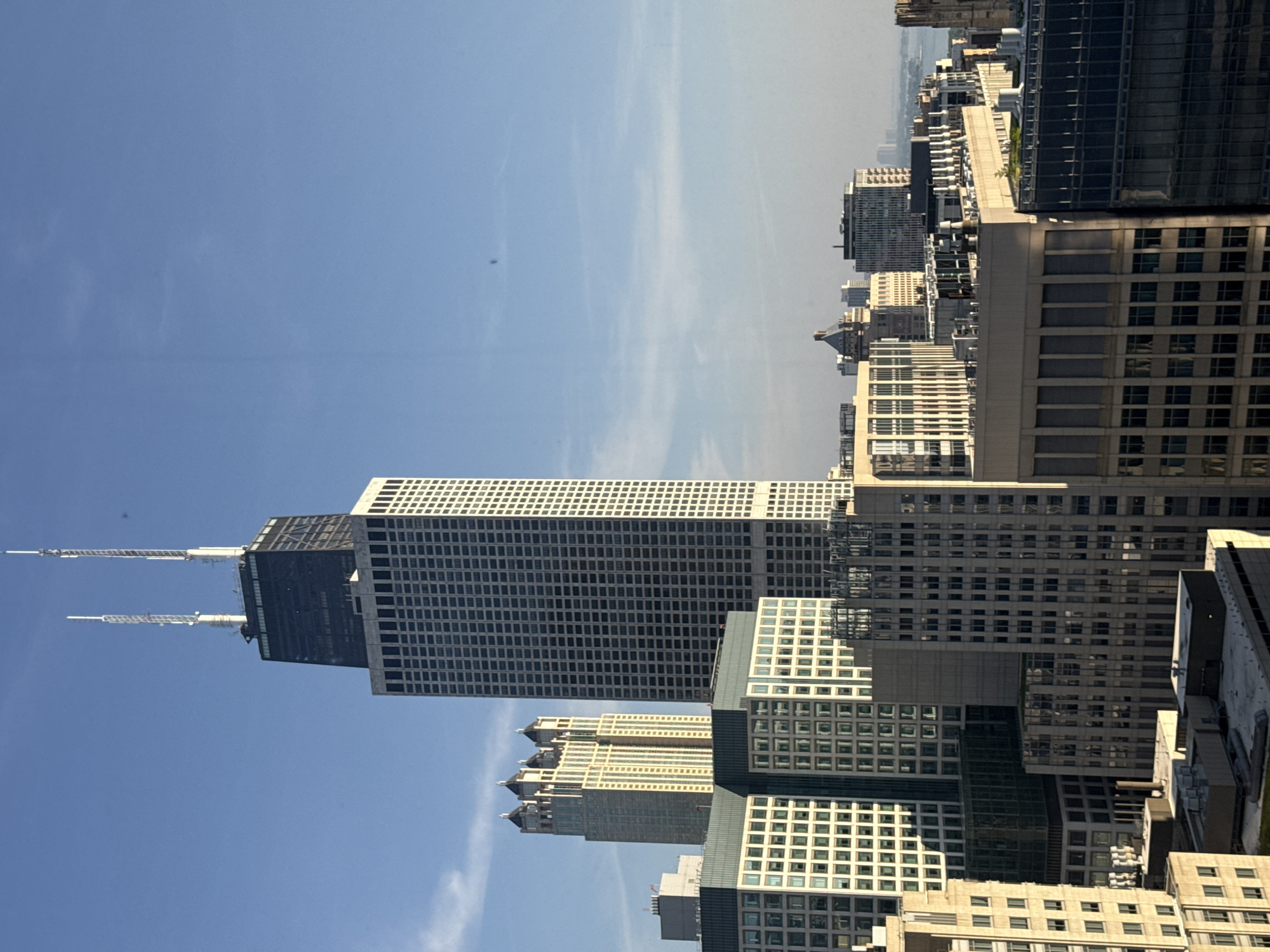 This is a photograph of the Chicago skyline, taken from a high vantage point, looking south-east. The most prominent building is the Willis Tower, formerly known as the Sears Tower, with its distinctive two antennas on top. The image captures a clear blue sky with some wispy clouds. In the mid-ground, several other skyscrapers and buildings are visible, showcasing a dense urban environment. The buildings vary in architectural style and height, with some featuring repetitive window patterns and others with more ornate designs. The lighting suggests it is daytime, likely morning or afternoon, with sunlight illuminating the eastern facades of the buildings. The overall impression is one of a vibrant and expansive cityscape.