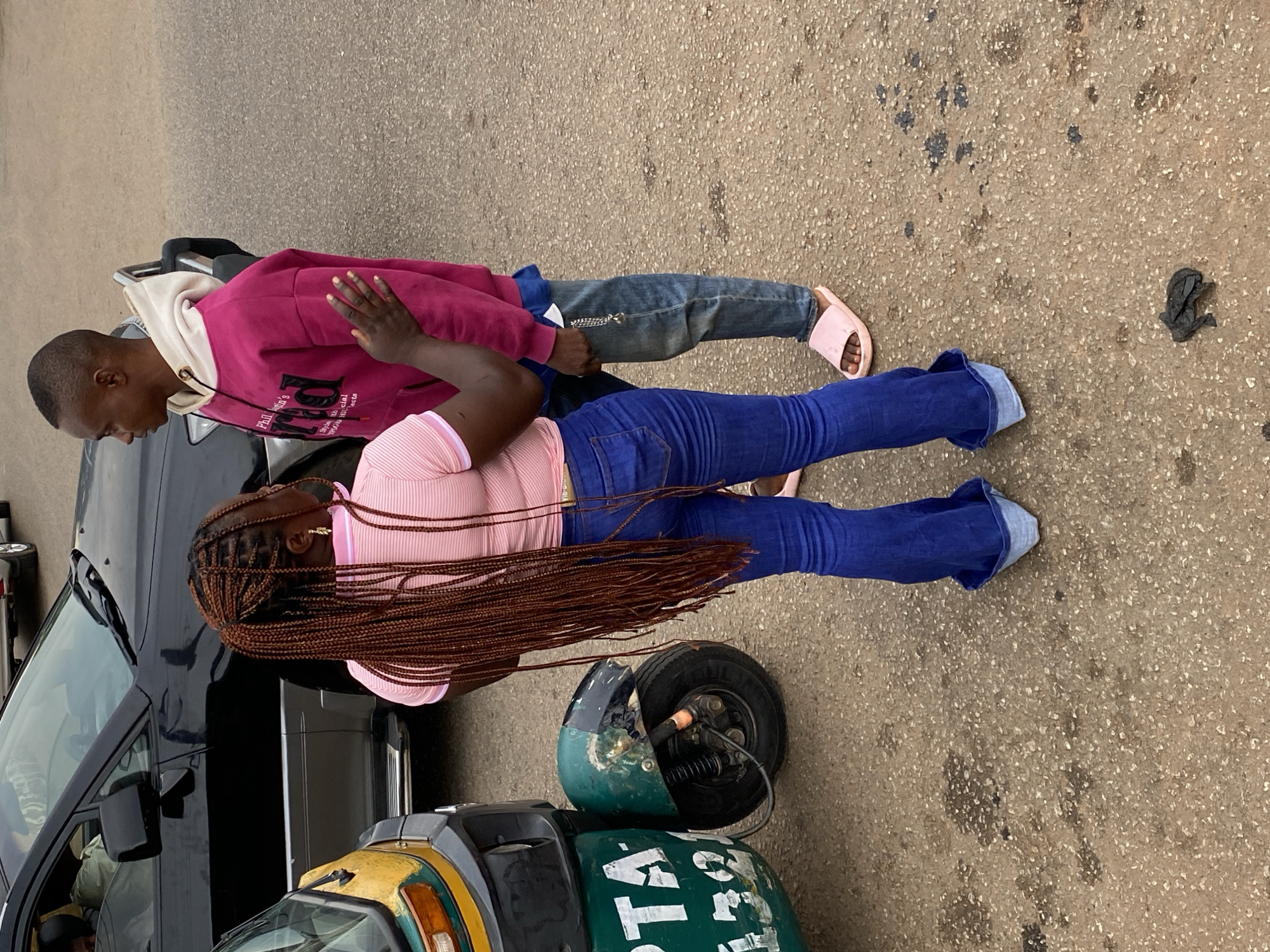 Two individuals are standing on a street in what appears to be Bukuru, Nigeria, based on the presence of a local "keke" or auto-rickshaw. The first individual is wearing a pink hoodie and jeans, with pink sandals. The second individual is wearing a pink striped top and blue jeans. Both appear to be standing near a black vehicle and the green "keke." A piece of litter rests on the street next to the individuals.