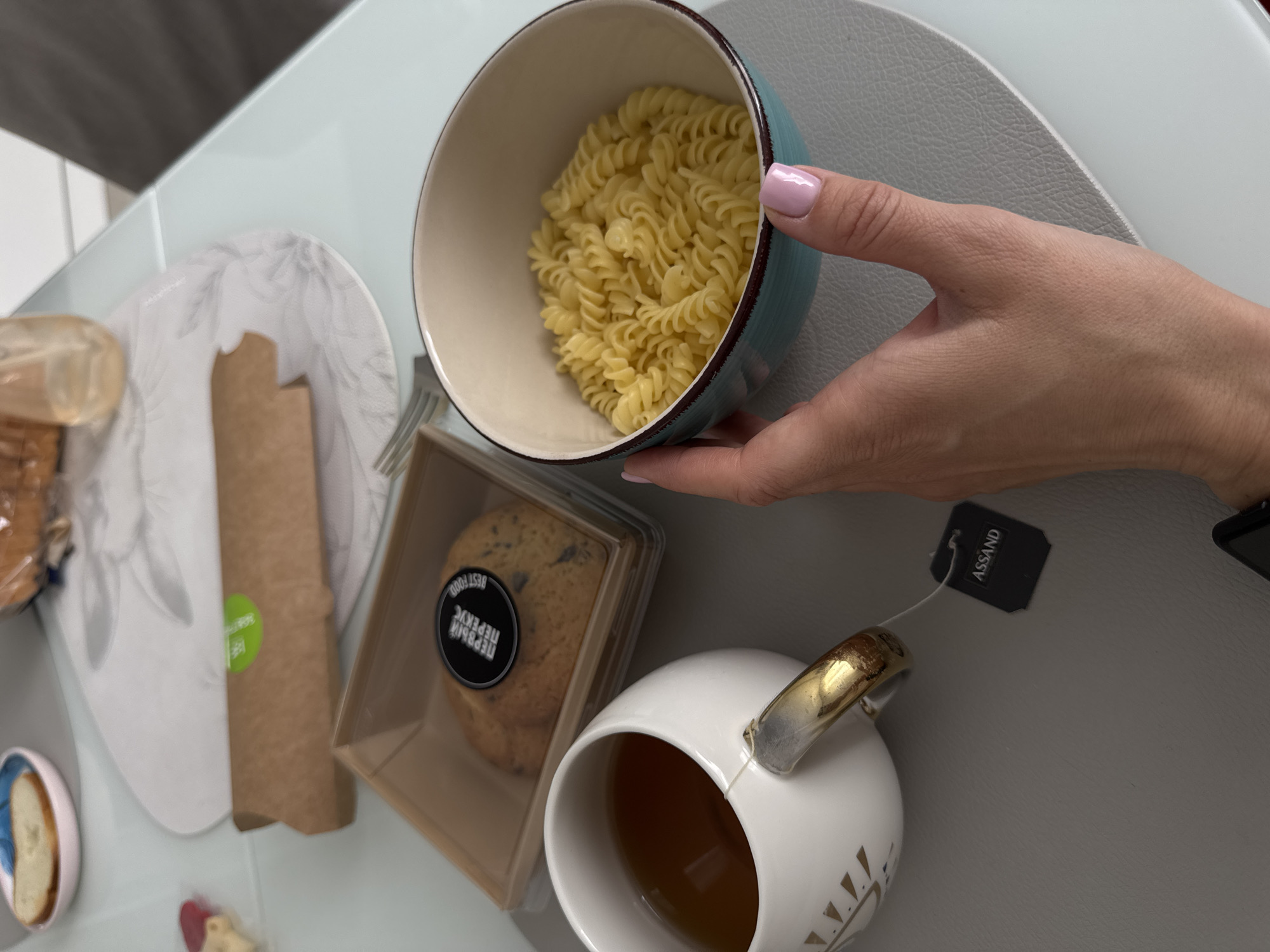 A high-angle shot presents a meal on a light grey surface. A woman's hand, adorned with a pink-polished fingernail, holds a blue-rimmed bowl filled with cooked rotini pasta. Beside the bowl, there's a plastic container holding a cookie with a circular label in a foreign language. Next to the cookie is a white cup of tea with a gold-colored handle, a black Assand tea bag tag hanging from it. Also, a brown packet with a green sticker on it is visible in the background. Based on these details and the presence of products labeled in Russian, the location is likely to be in Russia, possibly Sochi.