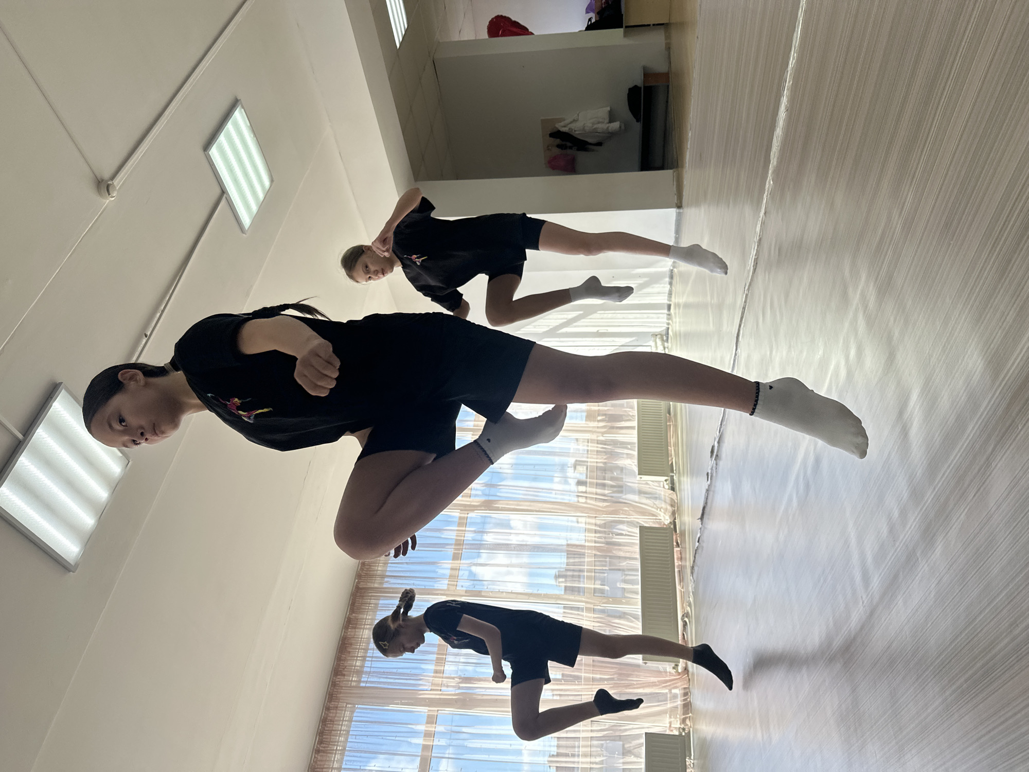 The image shows three young women in a dance studio in Votkinsk, Russia. They are dressed in black t-shirts and shorts, with various colored socks. All three are in dynamic poses, seemingly practicing a dance routine. The room has white walls, fluorescent lighting, and windows with drawn curtains. The floor is light-colored, possibly linoleum or a similar material. The perspective suggests the photo was taken from a low angle. There are also what appear to be mirrors in the background.