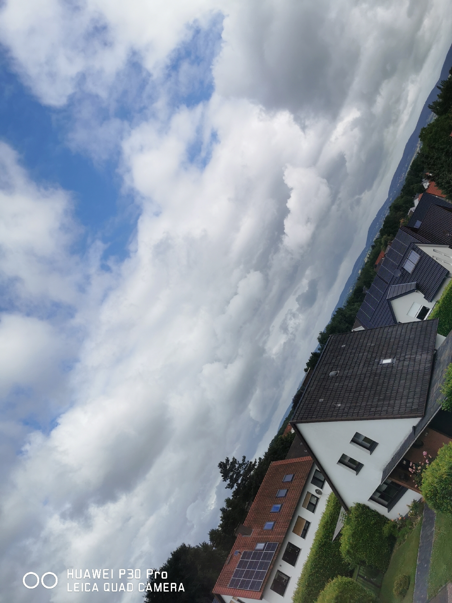 The image, captured with a Huawei P30 Pro camera, displays a suburban residential area with a cloudy sky. Several houses with tiled roofs are visible, some featuring solar panels. Lush green trees and manicured hedges surround the properties. A distant mountain range is discernible in the background, partially obscured by clouds. The composition is characterized by a diagonal tilt, with the sky occupying a significant portion of the frame. The timestamp and camera information, "HUAWEI P30 Pro LEICA QUAD CAMERA," are overlaid on the bottom left of the image. There are no people or animals present in the scene.