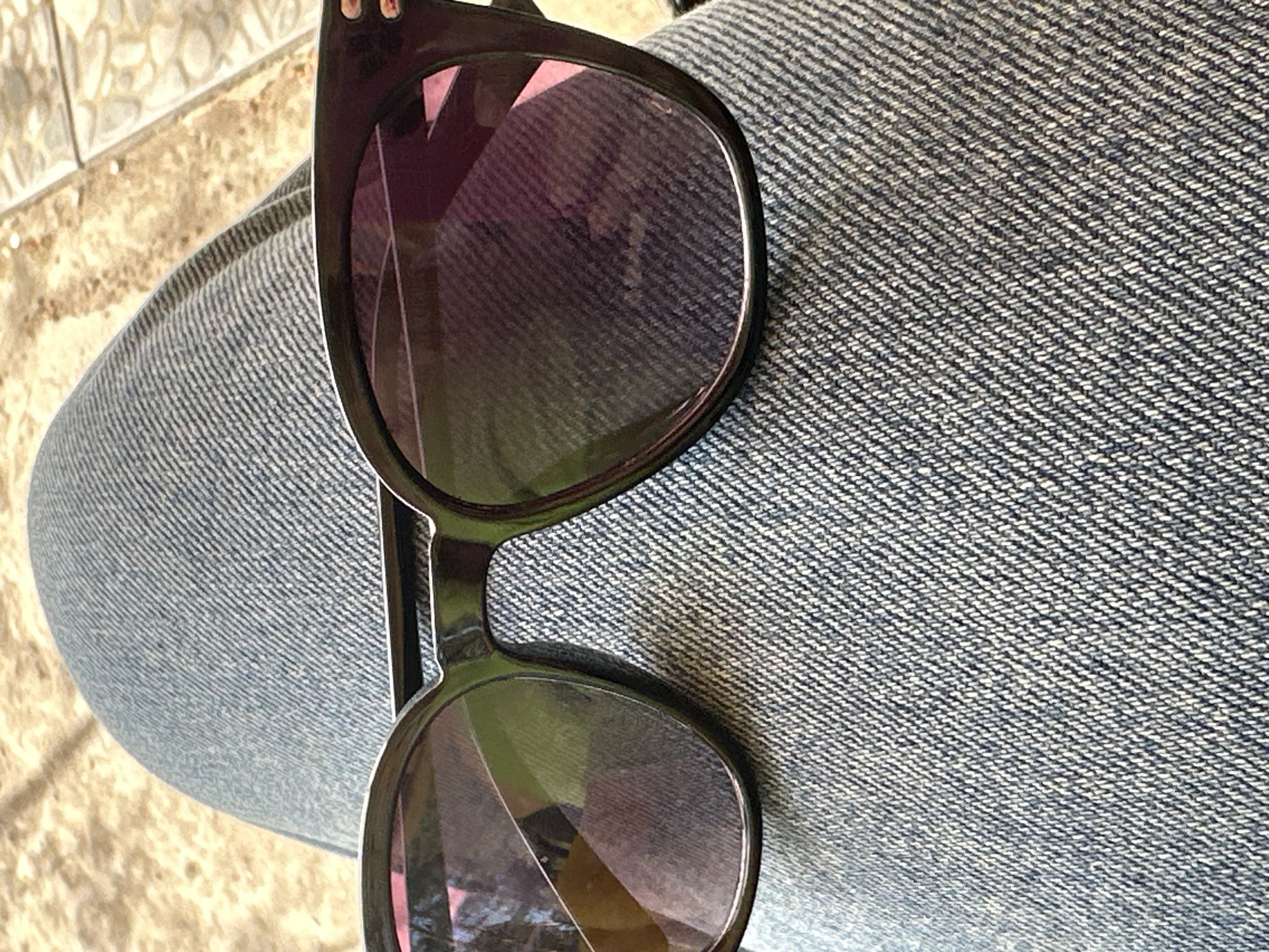 The image shows a pair of sunglasses resting on a person's lap. The person is wearing blue jeans. The background includes a glimpse of tiled flooring.