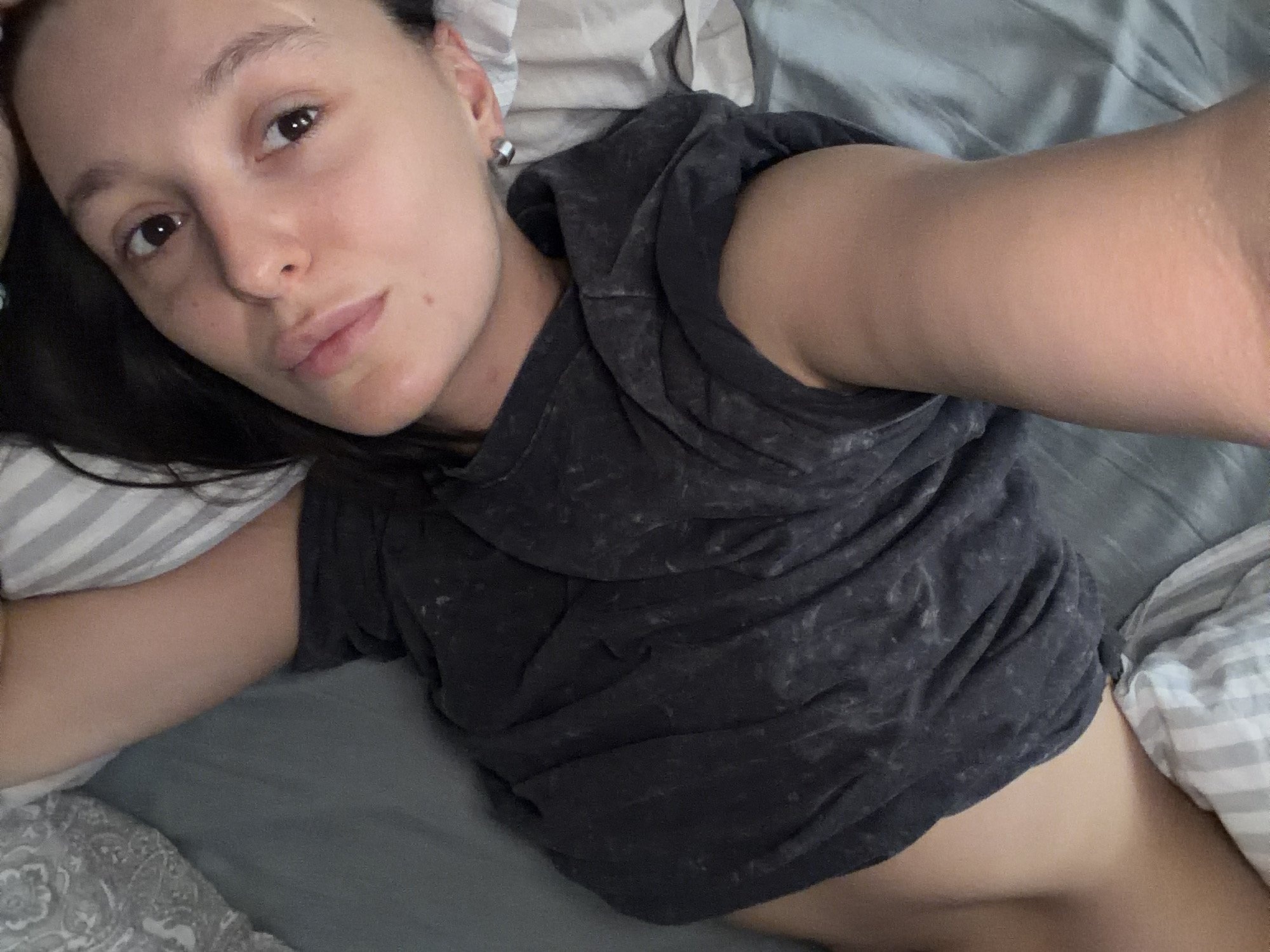 A young woman lies in bed. She has dark hair, light skin, and wears a dark gray t-shirt. The bedding is a mix of gray and striped patterns. The background is out of focus. There is no evidence to suggest the location is Moscow, Russia.