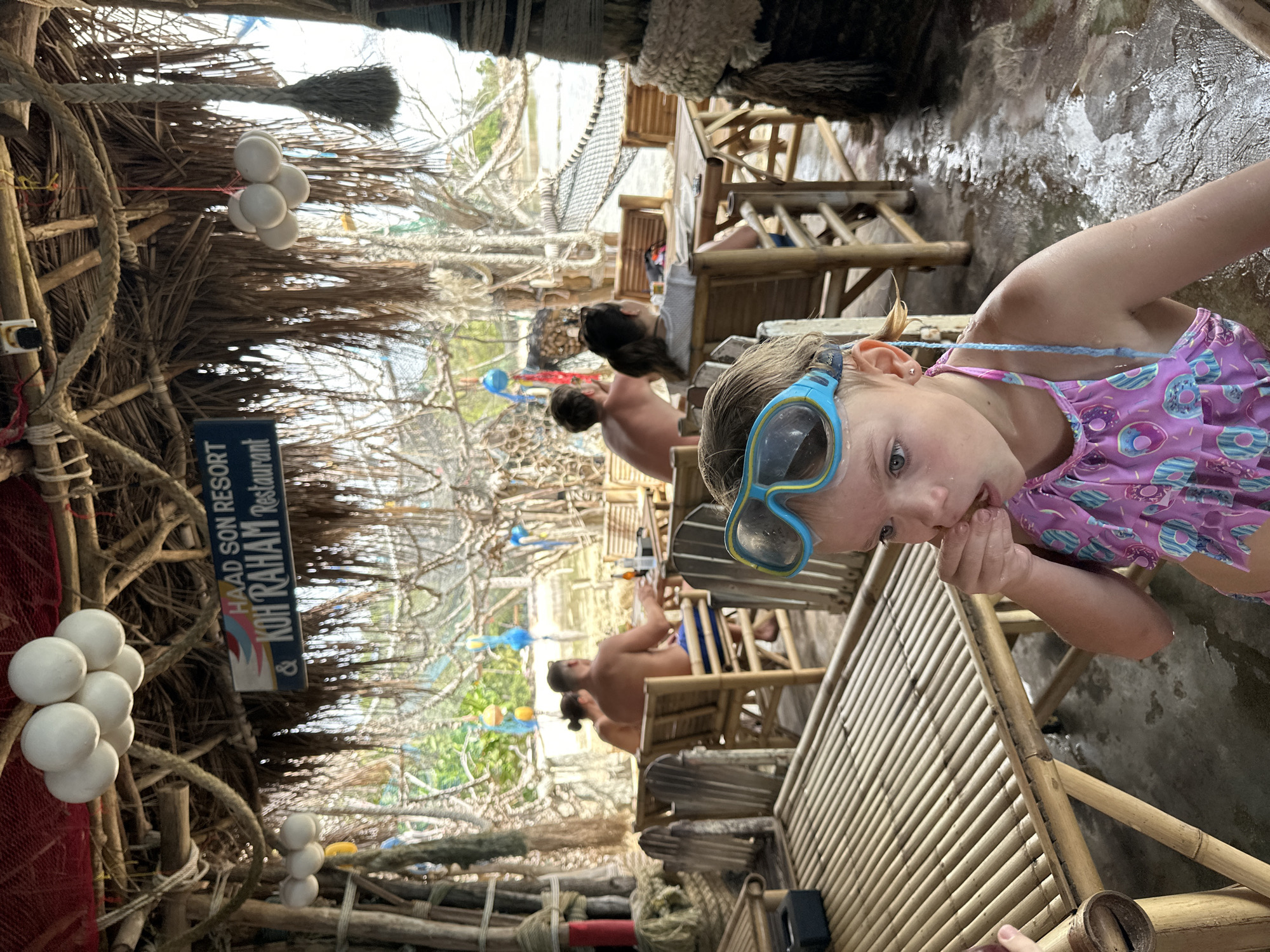 A young girl in a pink swimsuit with a donut pattern and blue goggles on her head sits in the foreground, eating. She is in a shaded outdoor restaurant with bamboo furniture and a thatched roof. Behind her, several people are seated at tables, with one couple appearing to be shirtless. The restaurant appears to be located on a beach or near water, with trees and foliage visible in the background. A sign reads "Haad Son Resort & Koh Raham Restaurant". The floor is wet and appears to be concrete.