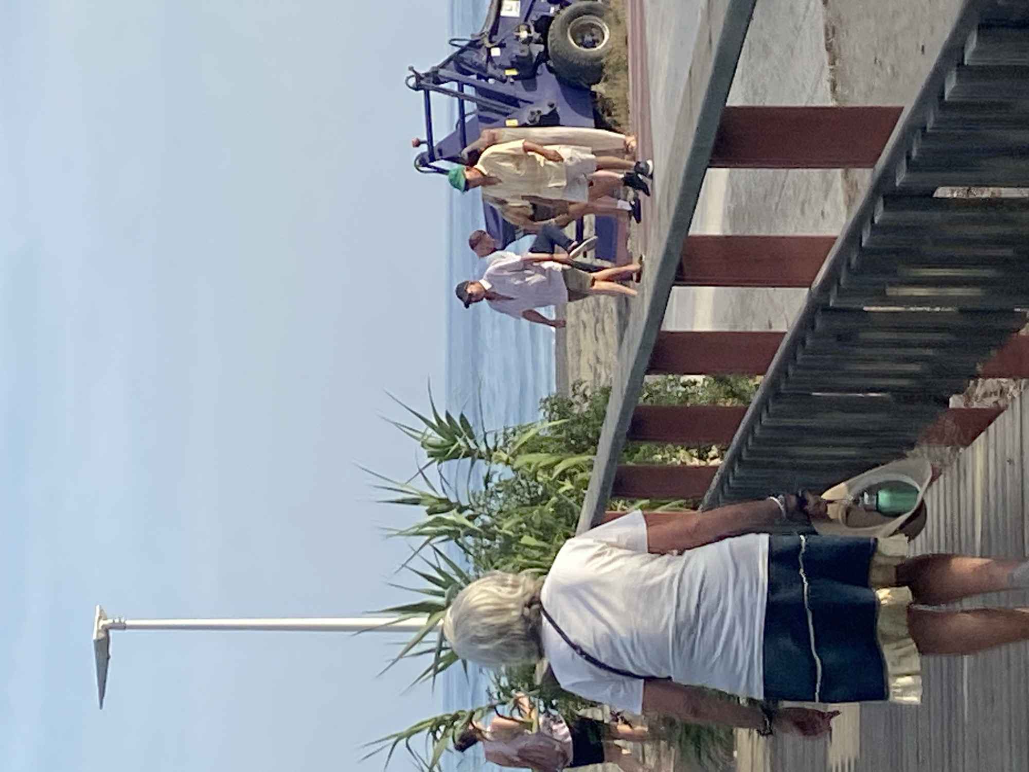 This media file depicts a scene on a sunny day, likely in Resinera-voladilla, Spain. The foreground features a person from behind, wearing a white t-shirt and blue shorts, holding a hat. In the midground, a group of people are gathered near a blue vehicle, possibly a tractor or a trailer, on a paved area next to a sandy beach and the ocean. The people appear to be casually dressed, in shorts and t-shirts, suggesting a relaxed, outdoor setting. The presence of the vehicle could indicate some sort of work or activity related to the beach or coastal area. The lighting suggests it is daytime. The overall atmosphere seems casual and warm, consistent with a beachside location. There is no visible text in the image.