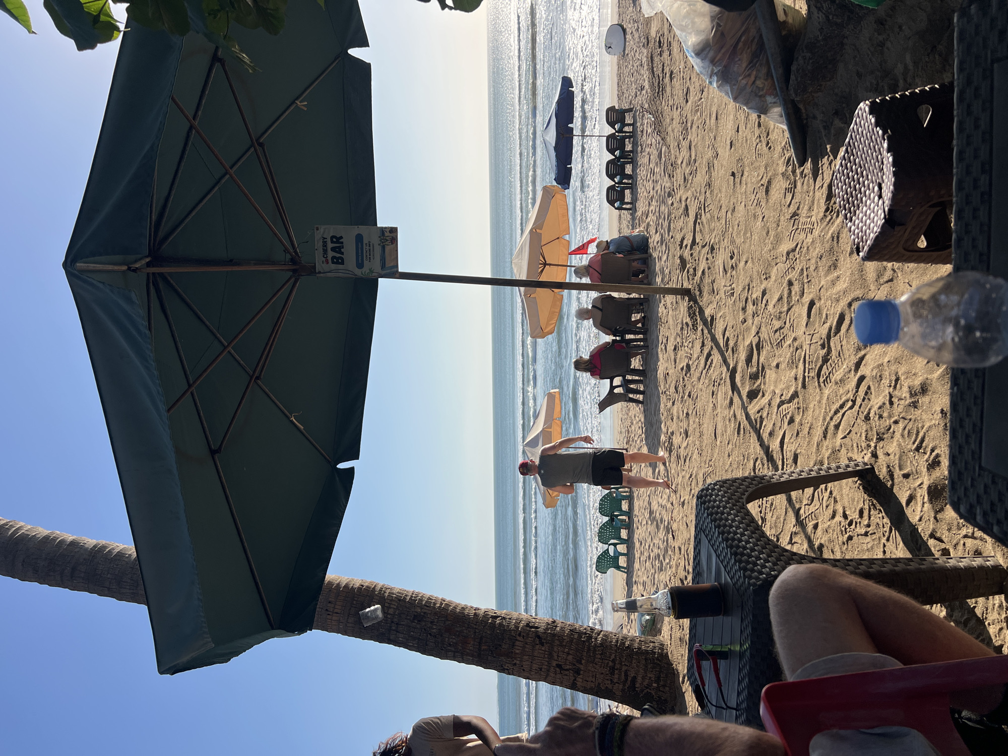 The image depicts a beach scene in Kuta, Indonesia. In the foreground, a table is set up on the sand, with a bottle of beer and sunglasses on it. A person's legs are visible, sitting in a red chair. Further back, a man in a dark t-shirt and shorts stands facing the ocean. Several beach chairs and umbrellas are set up on the sand, with some people seated under them. The ocean is visible in the background with gentle waves. A palm tree is on the left side of the image, and a sign for "CHERRY BAR" is hanging from an umbrella. The scene appears to be a relaxed setting at a beach bar.