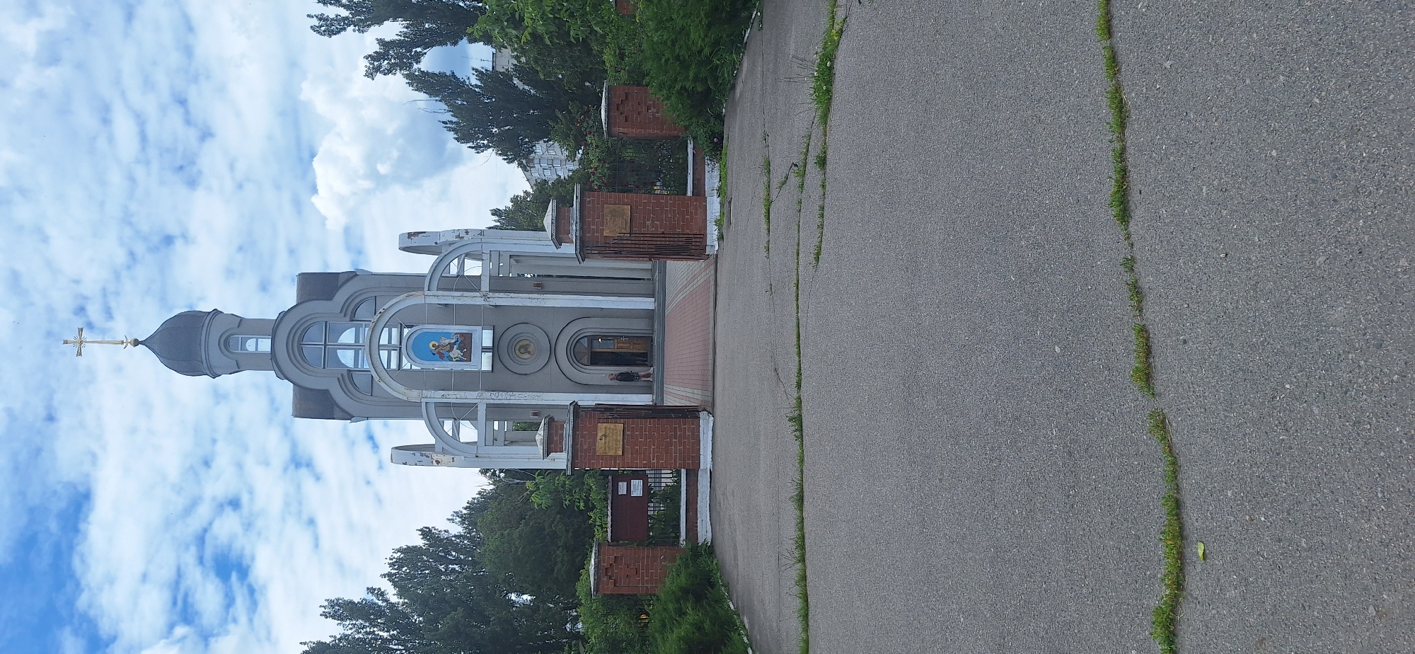 This is a vertical, eye-level shot of a light gray church building with a dark gray dome and a gold cross on top. The church has arched entrances with decorative white trim. A small, framed religious icon is visible above the main entrance. The building is situated behind a low, brick fence with brick pillars. A person, appearing as a child or small adult, is standing in the open doorway. The church is surrounded by green trees and foliage. The foreground is a cracked asphalt driveway with patches of grass growing through the cracks. In the background, a tall, multi-story residential building is visible above the trees. The sky is partly cloudy with white clouds against a blue background. The overall impression is of a religious building in an urban or suburban setting on a partly cloudy day. This is the St. George's Church, located at 30 Pushkina Street in Bila Tserkva, Ukraine.