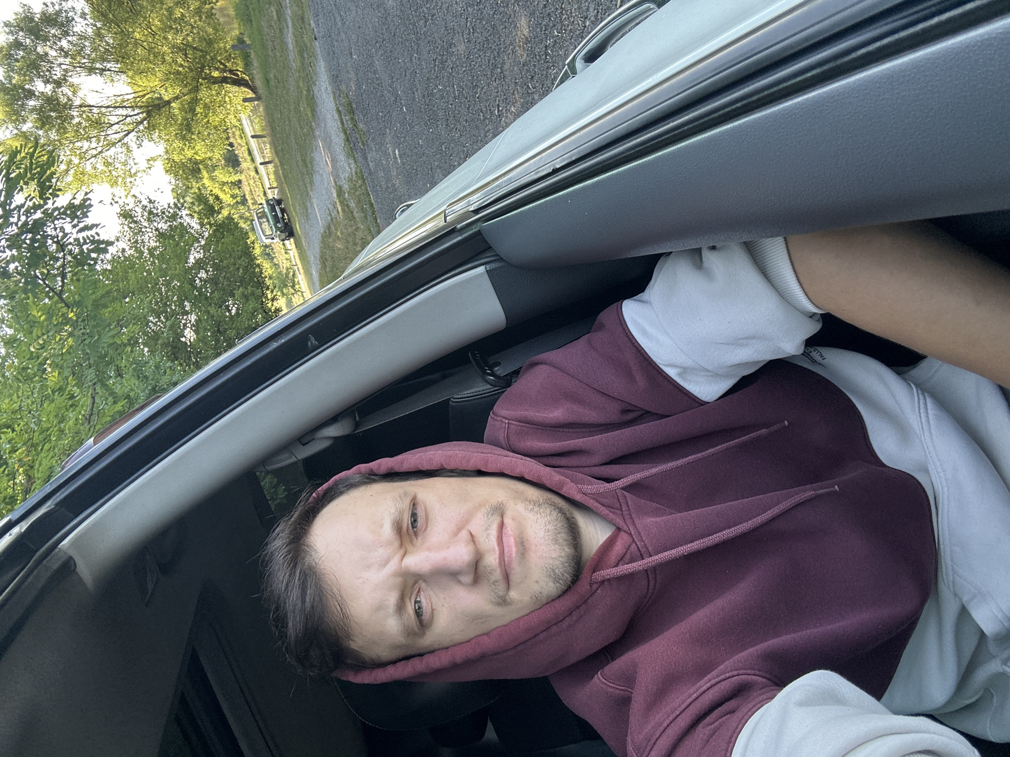 The image shows a man sitting in the driver's seat of a car, looking towards the camera. He is wearing a maroon and white hooded sweatshirt. The car is parked outdoors, with trees and a dirt road visible in the background. Another vehicle, a green jeep, is parked further down the road. The location appears to be a rural area, possibly a park or recreational area, outside of Makiv, Ukraine. The lighting suggests it is daytime, with sunlight filtering through the trees.