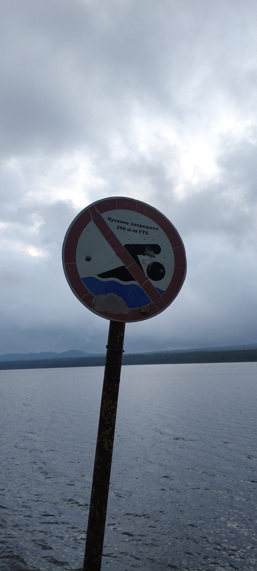 A weathered, circular sign with a red border, depicting a red diagonal prohibition line over a swimmer silhouette, is mounted on a rusty metal pole. The sign reads "Купание запрещено 250 м. от ГТС" (Swimming is prohibited 250 m from the dam). The sign is damaged with bullet holes and missing pieces. The pole is positioned on the shore of a large body of water, likely Lake Zyuratkul', with a distant, forested mountain range under a cloudy, overcast sky. The water is calm and reflects the muted light. The overall scene suggests a natural, possibly remote, location in Zyuratkul', Russia, with the sign indicating a restriction related to water safety.
