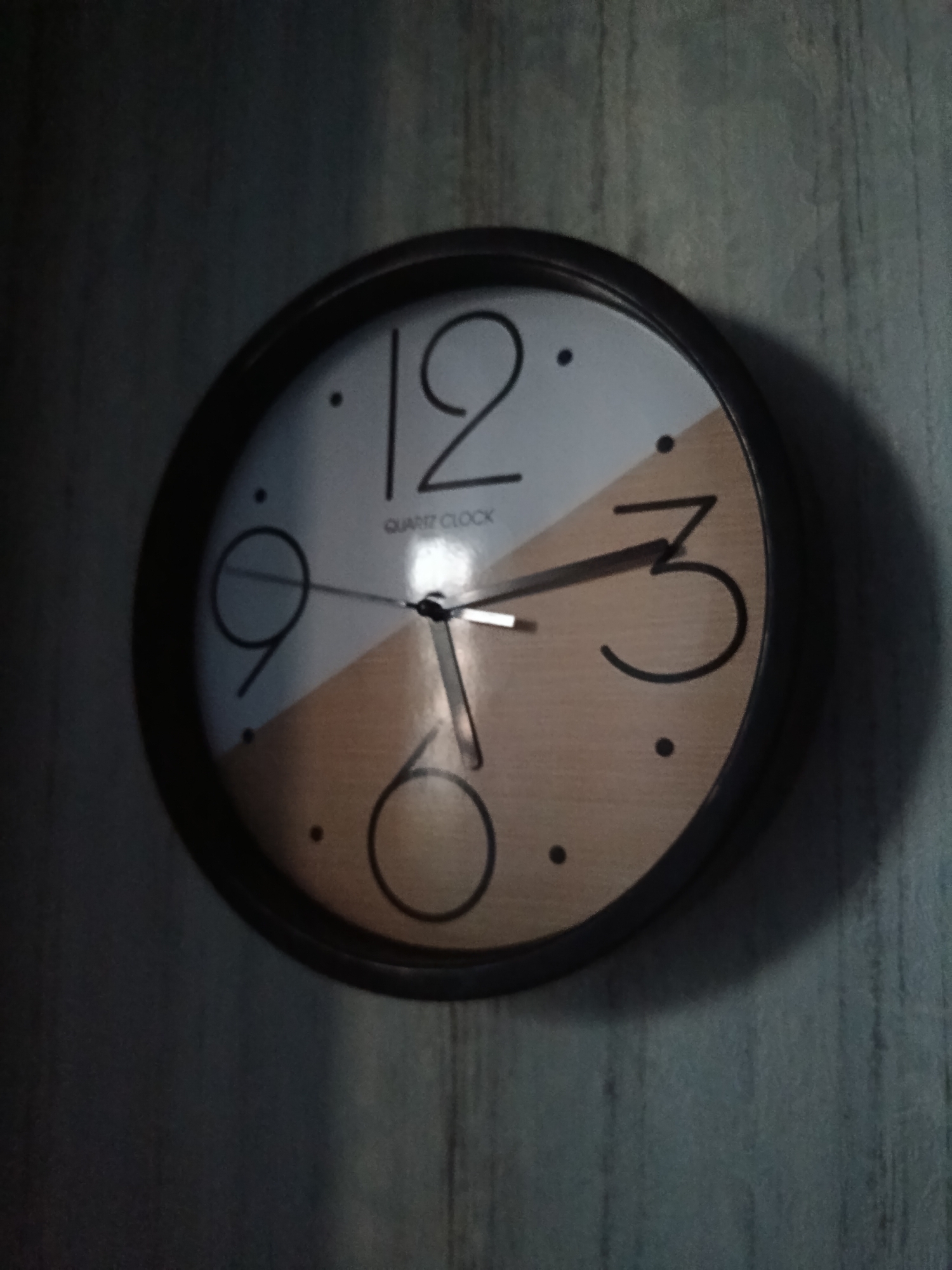An analog clock with a black frame hangs on a wall. The clock face is divided into two sections, one white and one a light wood grain. The numbers 12, 3, and 6 are visible, along with markings for other hours. The clock hands indicate a time between 6:30 and 6:45. The clock is positioned in the city of Cherkaske, Ukraine. The background appears to be a textured wall.