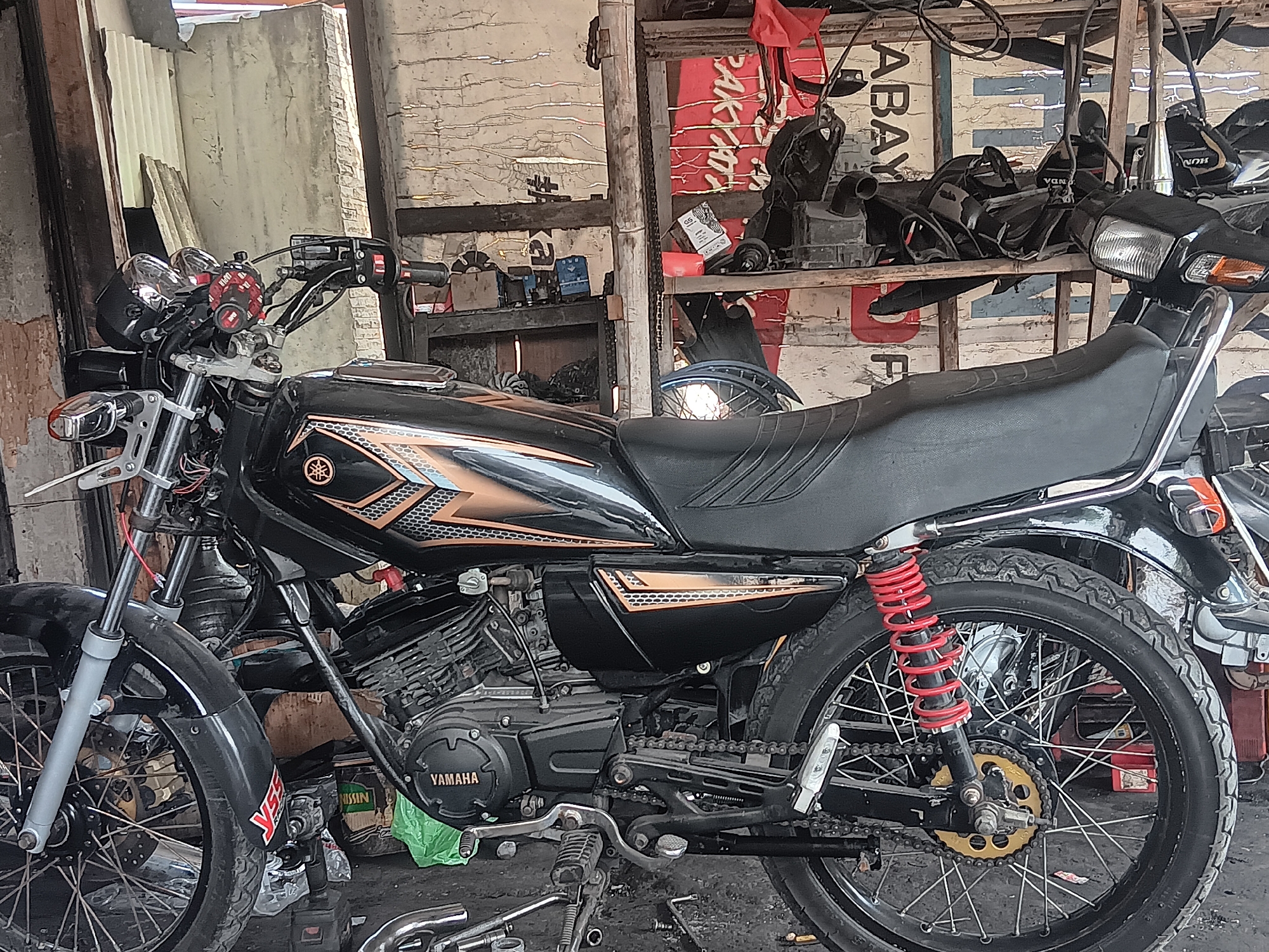 A black Yamaha motorcycle is parked inside a dimly lit garage or workshop in Batursari, Indonesia. The motorcycle is the primary focus, displaying its engine, chrome accents, and red rear shock absorbers. Various motorcycle parts, tools, and debris are scattered around the garage, suggesting ongoing repairs or storage. The background shows the textured walls of the workshop and shelves filled with additional parts, indicating a functional repair space. The lighting is subdued, with natural light filtering in from the entrance, creating a gritty and realistic atmosphere of a working garage.