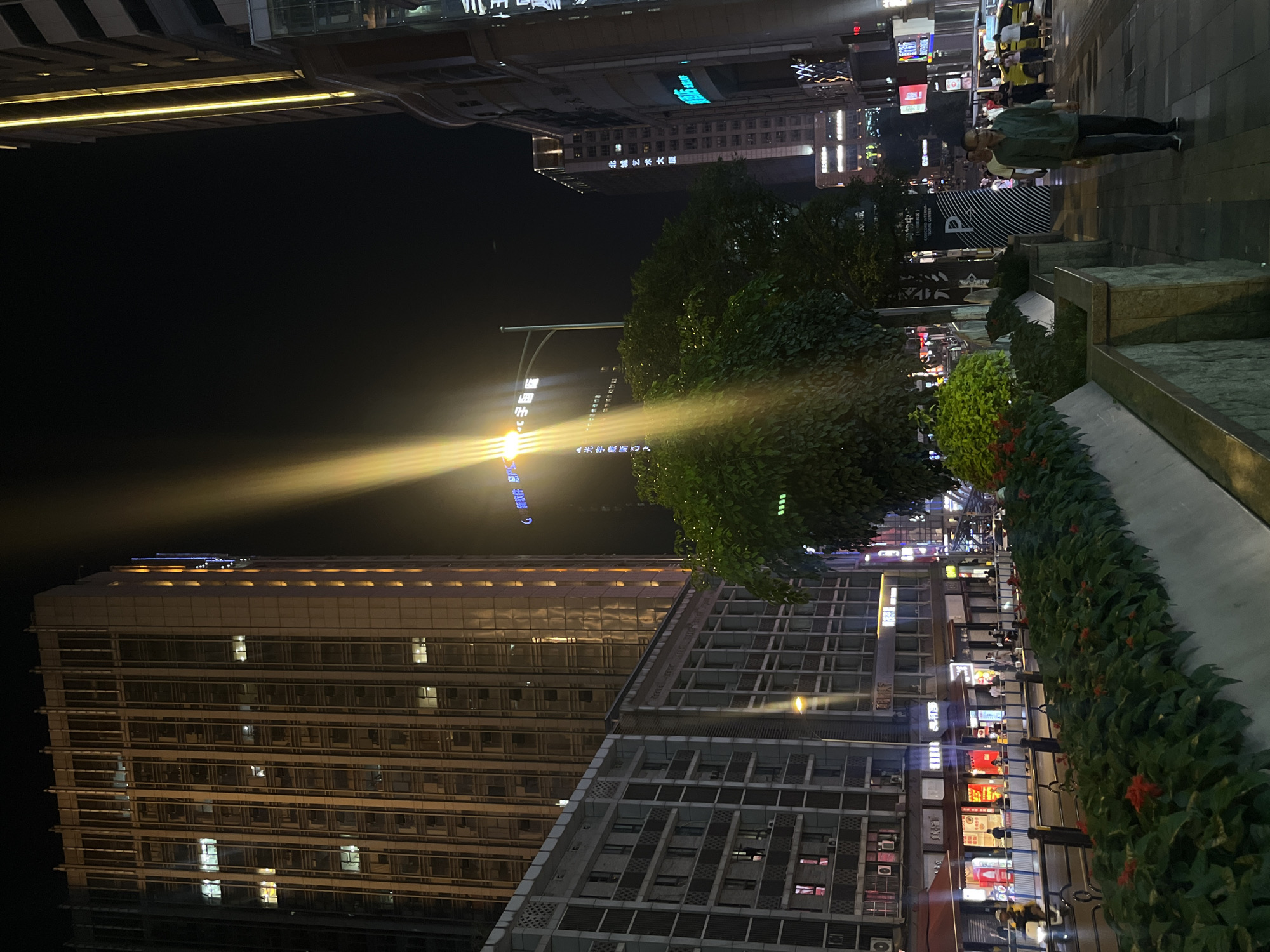 This nighttime image captures an urban streetscape in Liangjiang, China, characterized by illuminated buildings and artificial light. The sky is dark, highlighting the various light sources across the scene.
In the foreground on the right, a paved sidewalk is visible where at least two people are walking. The sidewalk is bordered by low planters filled with green bushes, some featuring small red flowers. Behind the bushes, shopfronts and building facades with glowing signs can be seen.
Tall, modern buildings dominate the background and left side of the image. One prominent high-rise on the left displays numerous illuminated windows, reflecting the surrounding light. Another building in the mid-ground features a bright, upward-projecting yellow-white light beam that cuts diagonally across the dark sky, creating a striking visual. More buildings with lit windows and signs are visible further back.
Visible Chinese text from signs and billboards includes "新软件总产", "宇国际", "▲光宇戴斷四", "北城艺", and "銀". The scene conveys a sense of a vibrant, active city environment after dark.