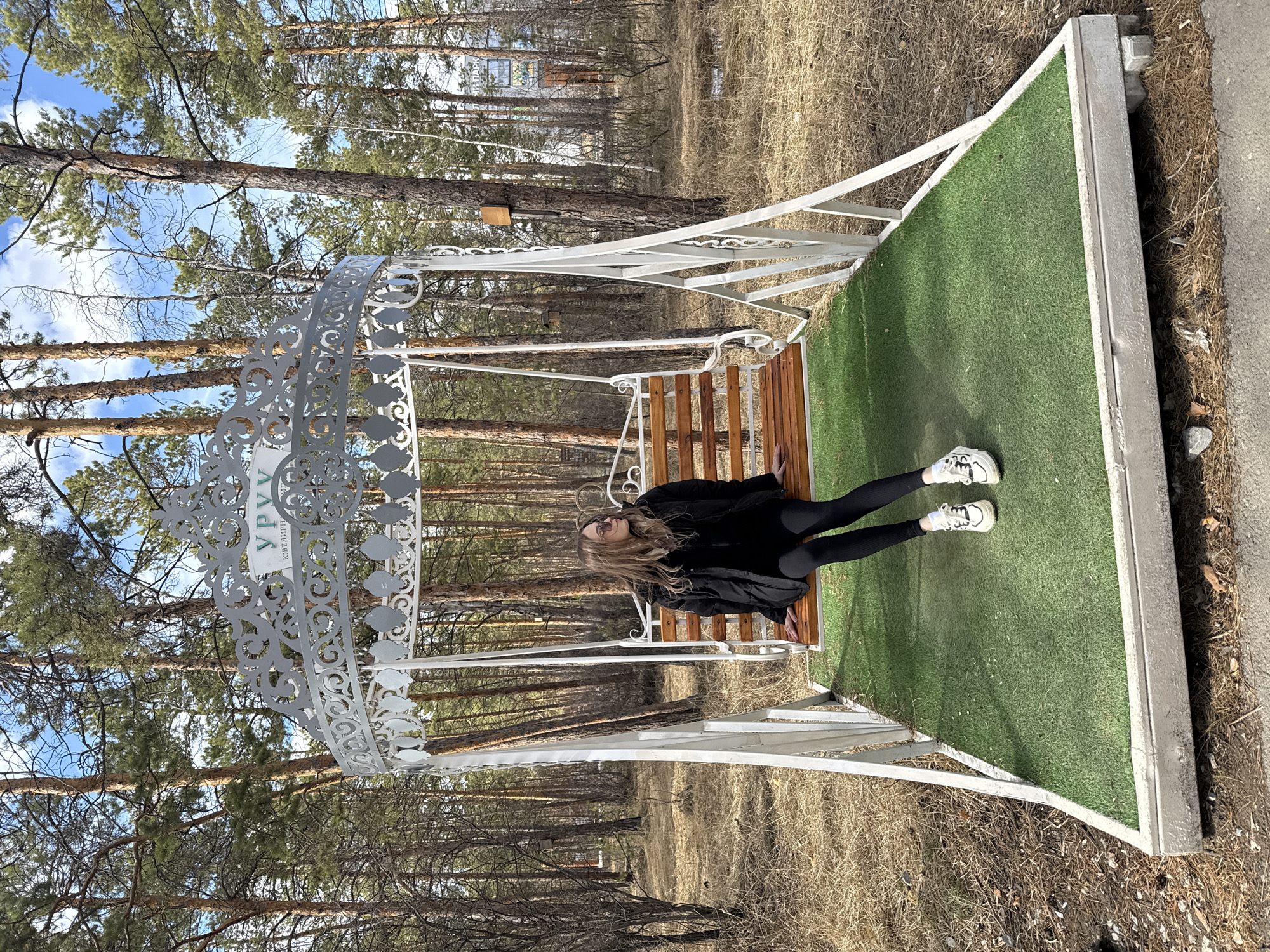 A blonde-haired woman wearing a black jacket, black leggings, and white sneakers lies on a wooden swing bench under a white, ornate metal canopy, in a wooded area in Yakutsk, Russia. The bench rests on a square platform covered with green artificial turf. The surrounding area consists of tall, slender pine trees with brown ground cover. The sky is blue with scattered clouds.