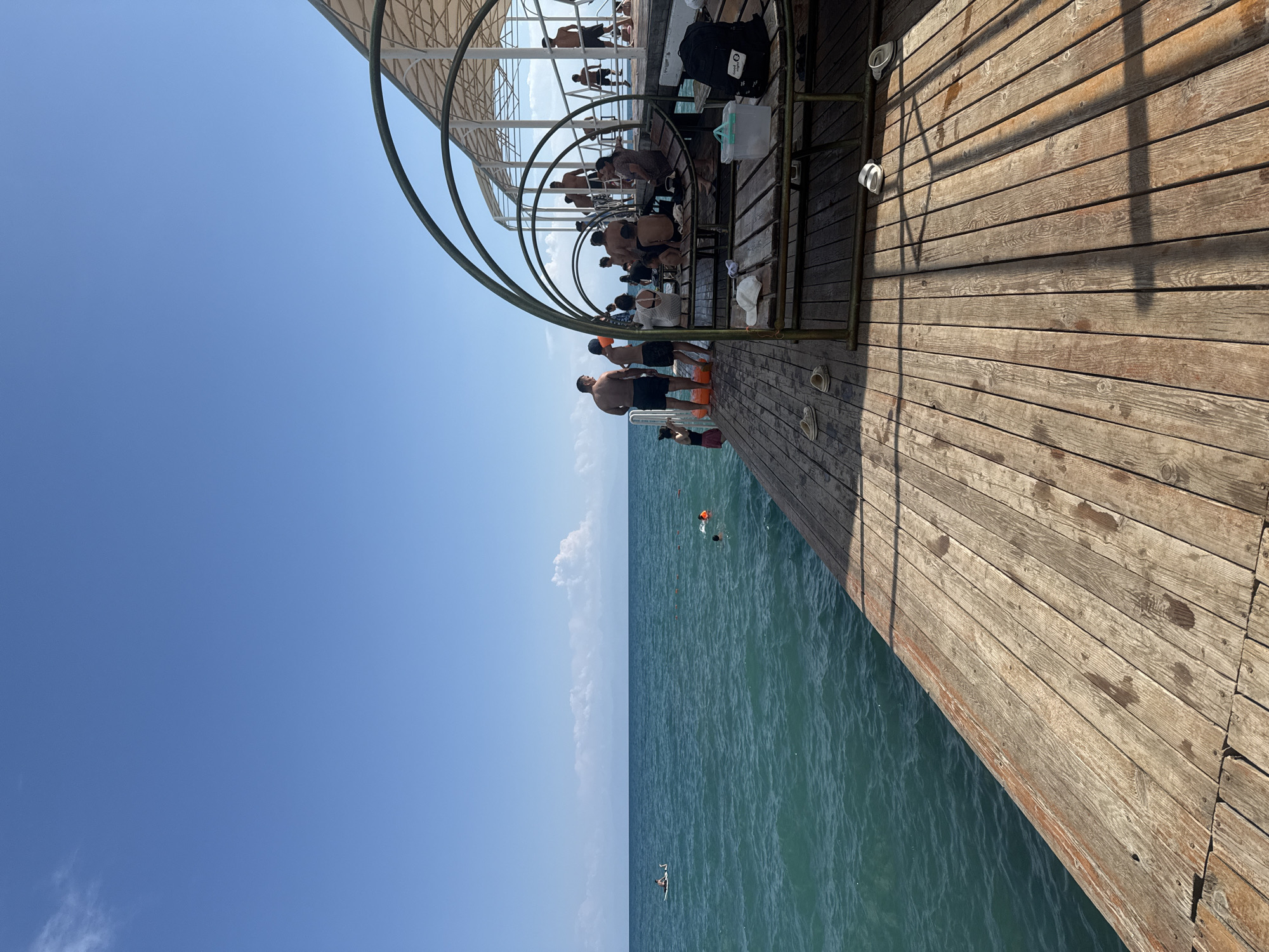 The image depicts a crowded wooden pier with people engaged in various activities. On the pier, individuals are standing, sitting on benches, and relaxing under a shaded structure with a white canopy and arched supports. Some people are dressed in swimwear. Scattered on the wooden planks are several pairs of sandals and flip-flops, a white hat, and a black backpack.

Adjacent to the pier, the turquoise waters of a lake are visible, with ripples on the surface. In the distance, several people are in the water; one person is on a paddleboard, and others are swimming, with at least one wearing an orange life vest. The horizon is marked by a faint line of distant land or mountains under a clear blue sky with a few wispy clouds. The scene is consistent with a public beach or recreational area, likely located in Sary-Oy, Kyrgyzstan, given the context provided. The sun is casting shadows on the wooden pier, indicating it is daytime.