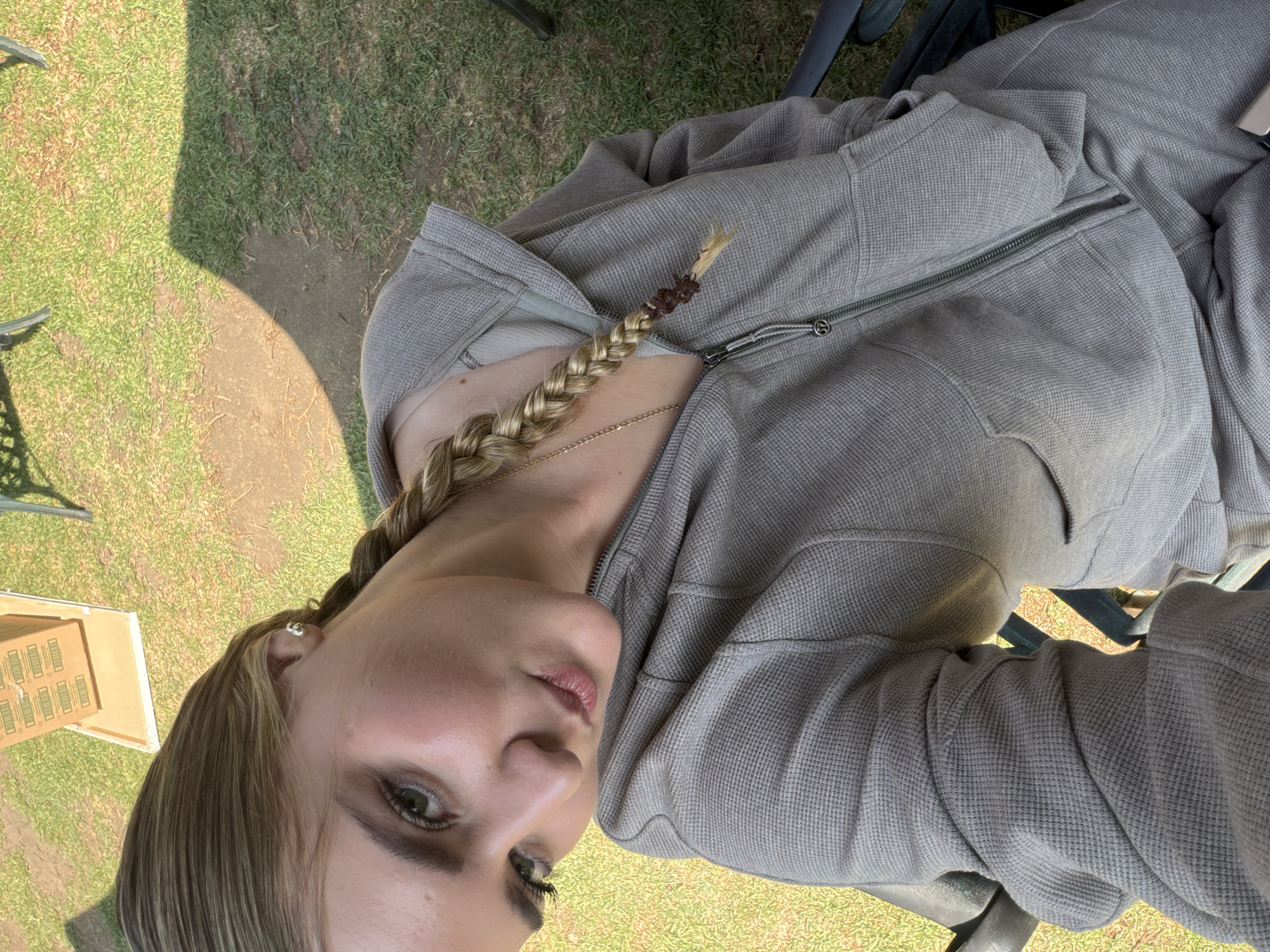 The image shows a light-skinned person with blonde hair braided on the left side of the frame. The braid is secured with a dark brown tie near the end. The person is wearing a gray hooded sweatshirt or jacket, zipped up to the chest. A thin gold necklace is visible. The person is looking towards the lower left of the frame.
The background shows a grassy area with patches of bare earth. A small wooden structure is visible in the lower left corner, possibly a table or box. Portions of chairs are also visible, suggesting an outdoor seating area.
Based on the request, the location is San Andrés Cholula, Mexico, however, no explicit details in the image confirm the location.