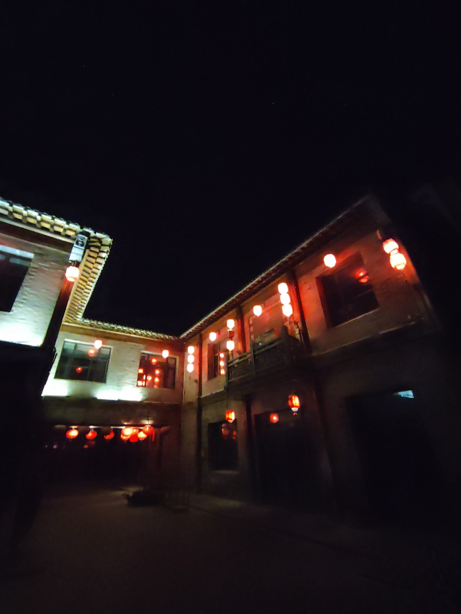 The image depicts an outdoor scene at night in what is identified as Lindian, China. The setting appears to be an alleyway or courtyard surrounded by traditional Chinese-style buildings. The architecture features tiled roofs and brick walls, with illuminated windows. The primary illumination comes from numerous red lanterns hung throughout the area. These lanterns cast a warm glow on the buildings and the ground, creating a festive and atmospheric ambiance. The sky above is dark, with a few faint stars visible. There are no people or animals present in the image. The ground appears to be paved or made of dirt. The overall impression is of a quiet, culturally significant location illuminated for nighttime viewing.