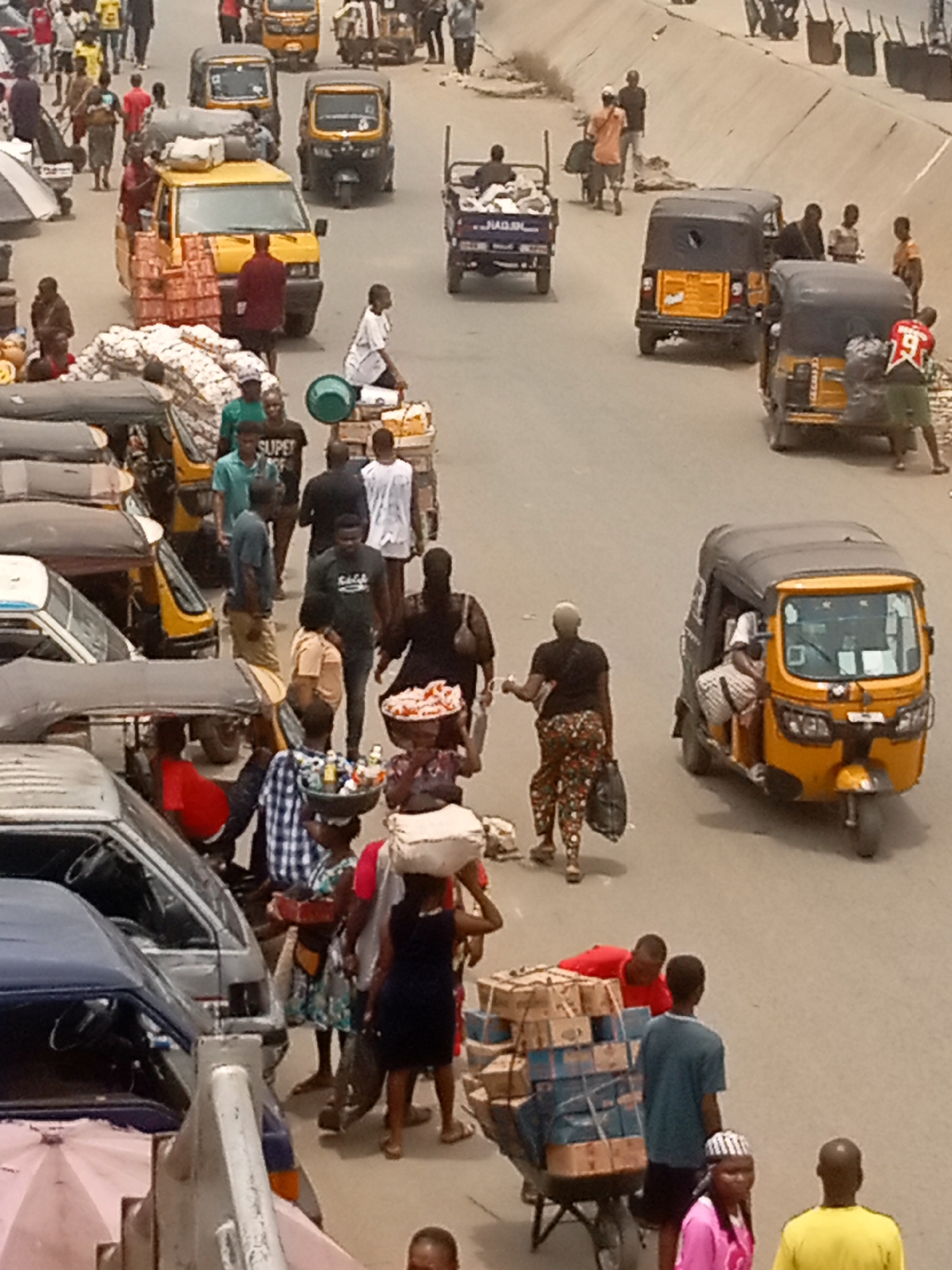 The image depicts a busy street scene in Onitsha, Nigeria. Several "keke" (auto rickshaws) and a few vans are visible, along with numerous pedestrians. Many people are carrying items on their heads or pushing carts loaded with goods. Some individuals appear to be engaging in commerce, while others are simply moving through the area. The environment suggests a bustling market or commercial district.